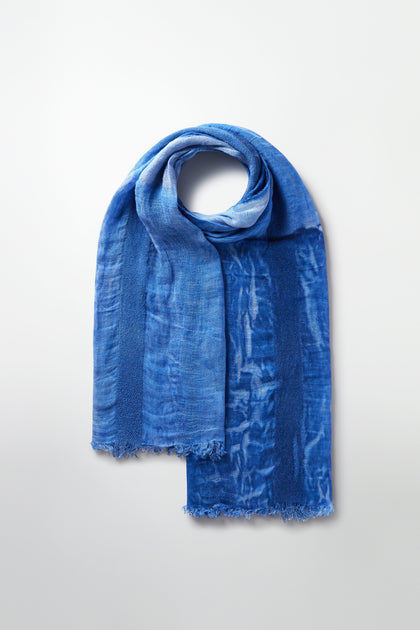 The Sky Linen Mix Scarf, crafted with linen in varying blue shades and a subtle pattern, is neatly folded on a plain background. Frayed edges add character to this elegant piece.