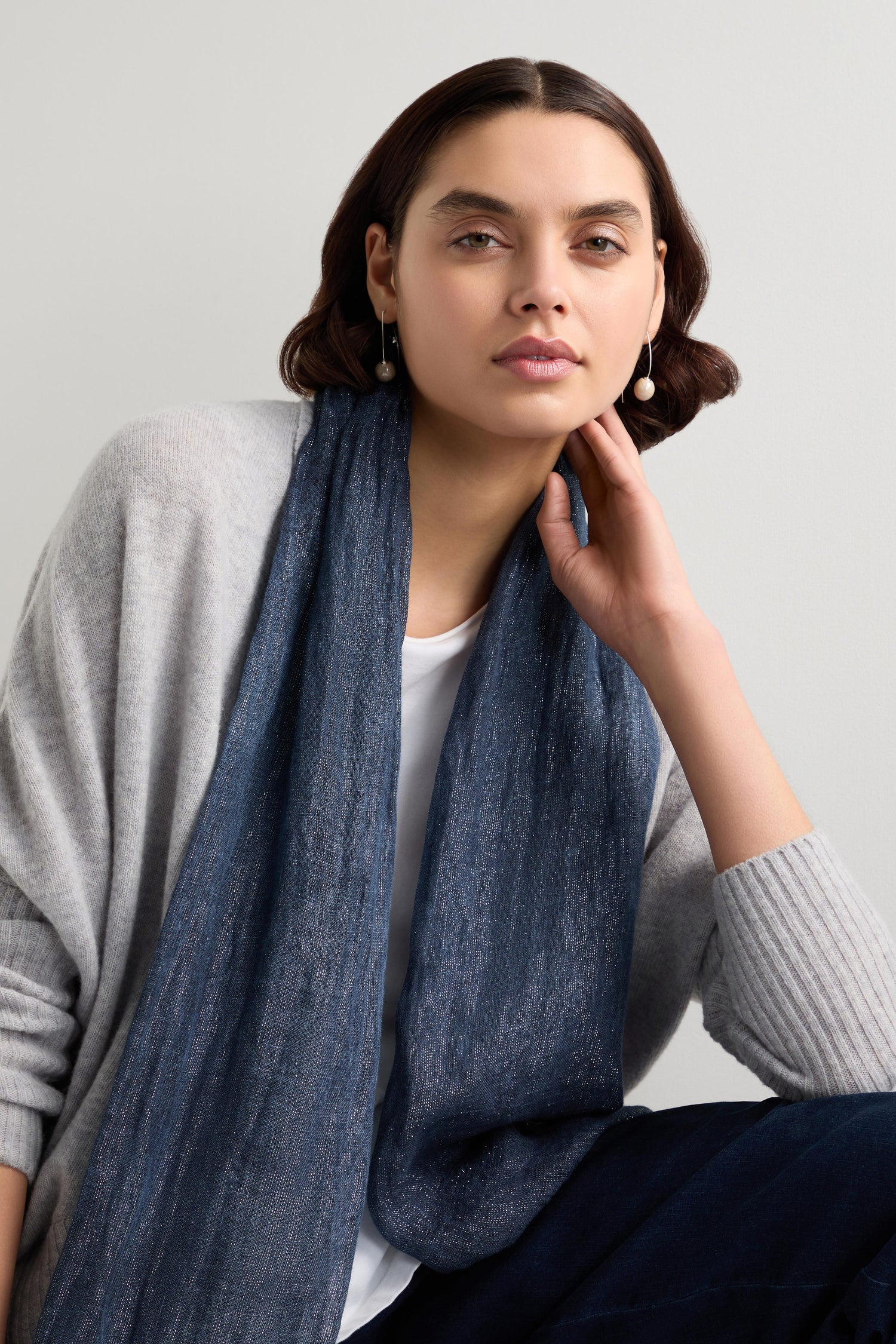 A person is seated, donning a gray sweater over a white shirt with the Starry Night Scarf, an indigo blue piece threaded with metallic highlights. They rest their head on their hand, gazing ahead.