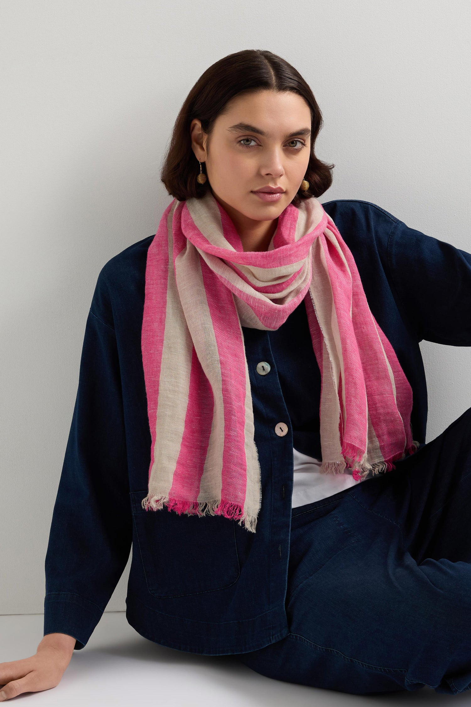 Person with shoulder-length hair in a blue outfit, accessorized with the Linen Stripe Scarf featuring fuchsia stripes, seated against a plain background.