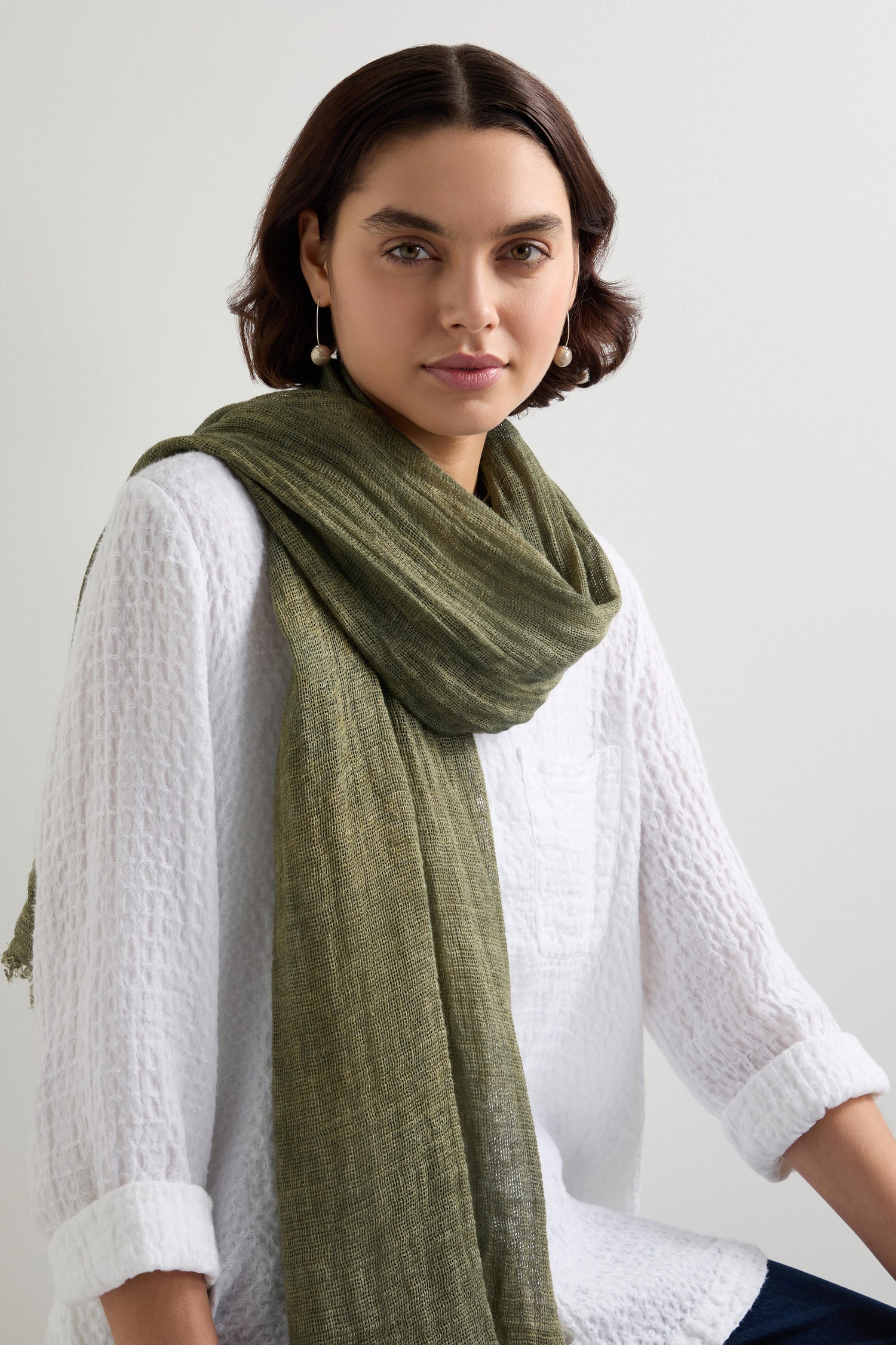 Someone with short hair wears a textured natural fiber white top paired with an olive green Woven Linen Scarf. The raw-edge finish adds rustic elegance against the neutral background.