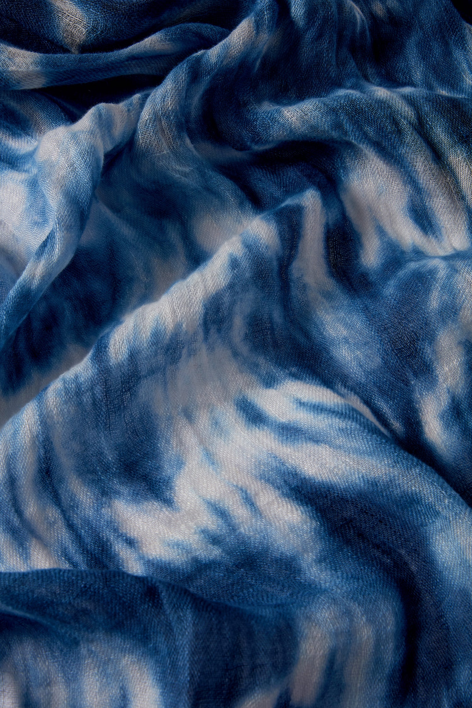 The Tie Dye Linen Scarf in indigo blue showcases a captivating crinkled fabric with swirling tie-dye patterns, ideal for adding bohemian flair to your summer wardrobe.