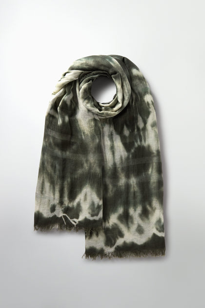 The Sparkle Tie Dye Linen Mix Scarf features an elegant circular drape in a luxurious linen-cotton blend. Its dark green and cream hues, enhanced with touches of khaki green, create a serene yet striking visual against the plain background.