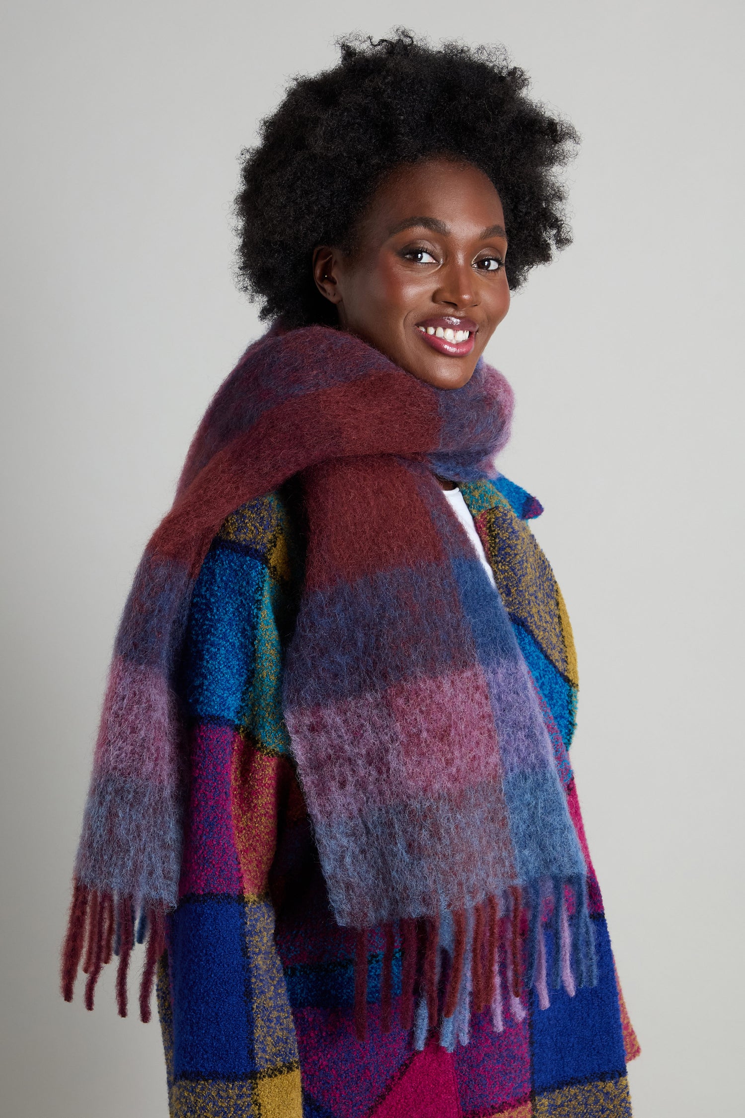 A person wears a vibrant patchwork coat and the Check Tassel Scarf, crafted from a mohair wool blend, smiling at the camera against a neutral background.