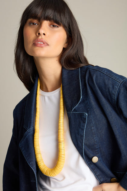 A person with long dark hair and bangs wears a denim jacket, white shirt, and a Long Graduated Coco Disc Necklace with yellow graduated disc beads, looking directly at the camera.