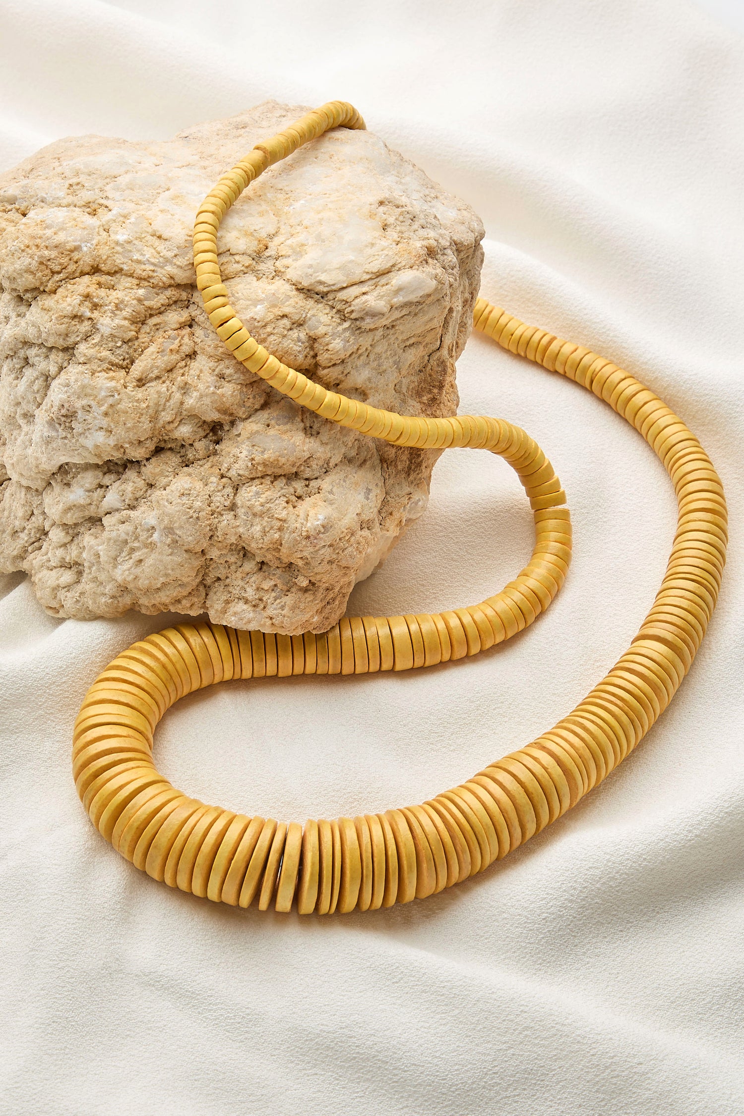 The Long Graduated Coco Disc Necklace, featuring meticulously crafted graduated disc beads by skilled artisans, is elegantly draped over a textured light-colored rock on a soft white fabric background.