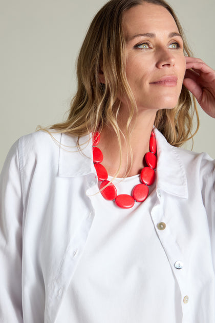 A person with long hair wearing a white shirt and the Short Wooden Pebble Necklace, looking off to the side.