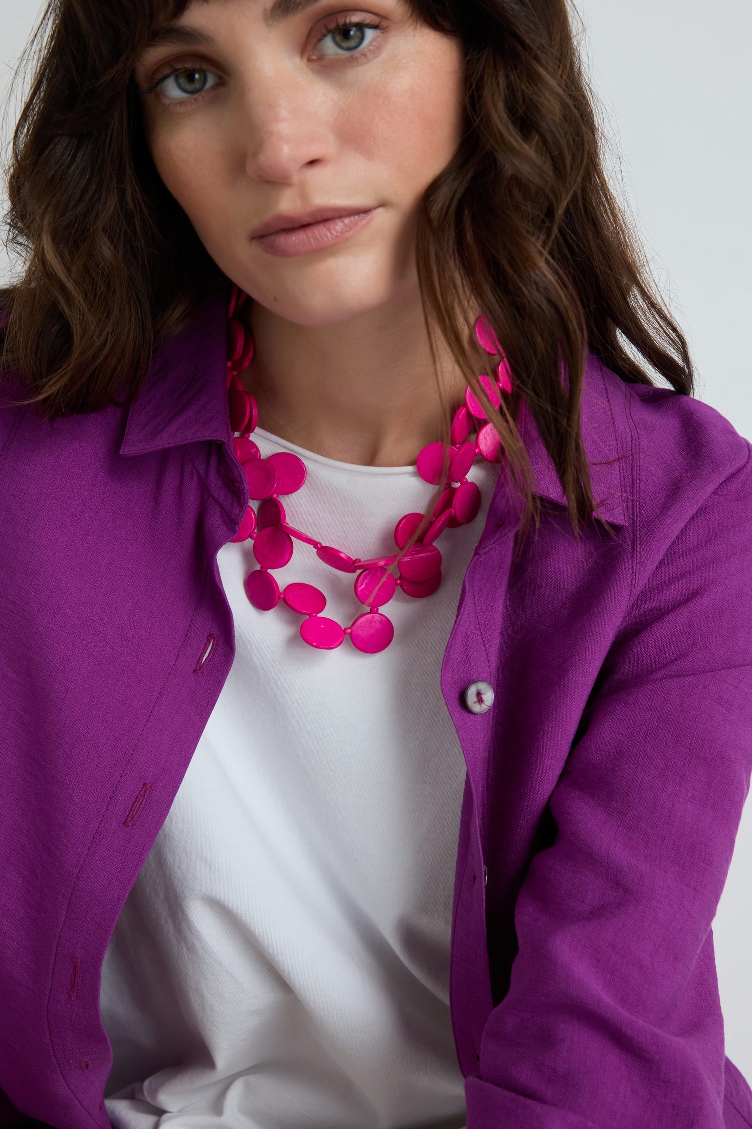Woman with wavy brown hair wears a white shirt, purple jacket, and a bright pink Long Strand Coco Disc Necklace handcrafted from coconut shell jewelry, looking at the camera against a plain background.