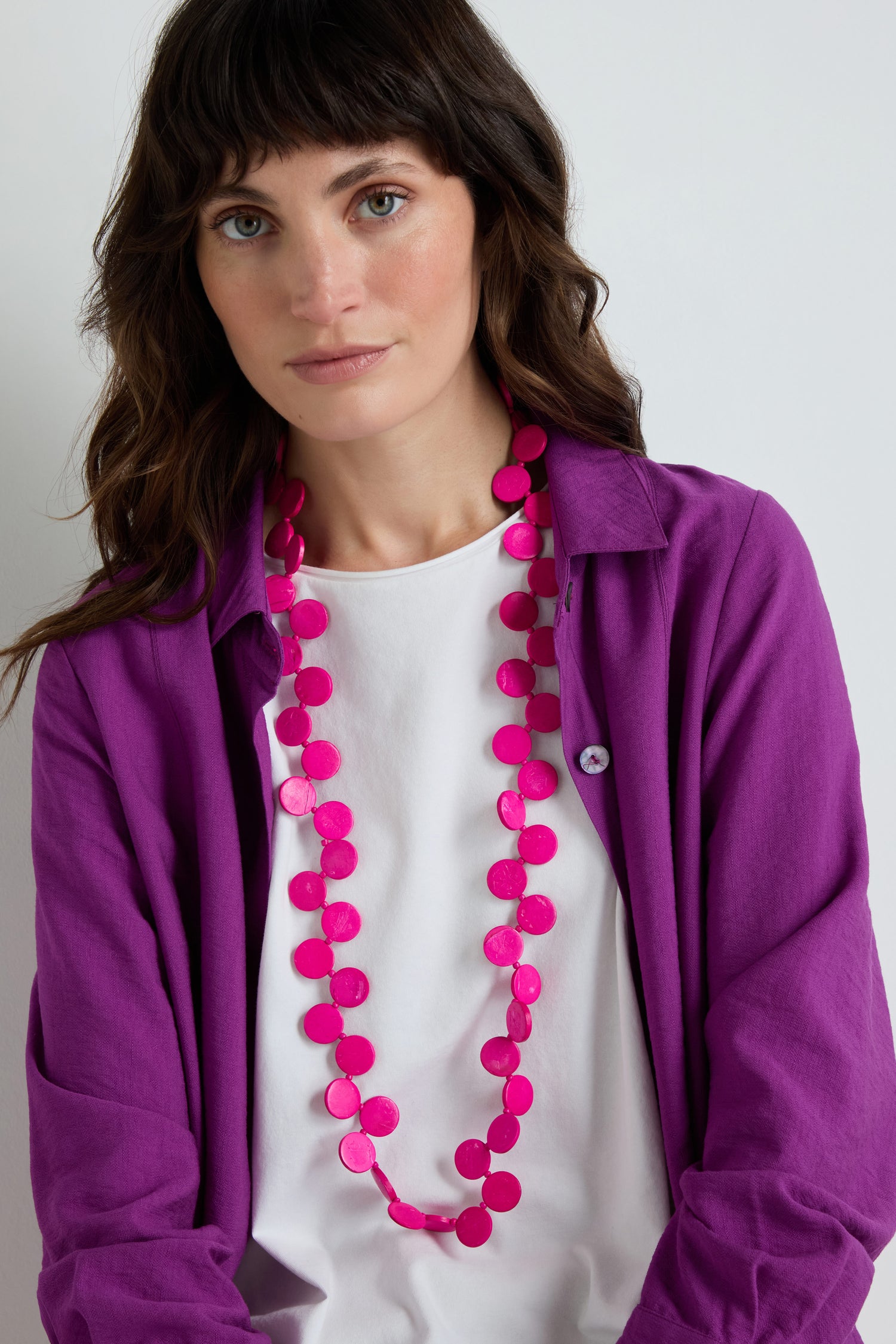 A woman with wavy brown hair poses in front of a plain white background wearing a white t-shirt, a purple button-up shirt, and the Long Strand Coco Disc Necklace.