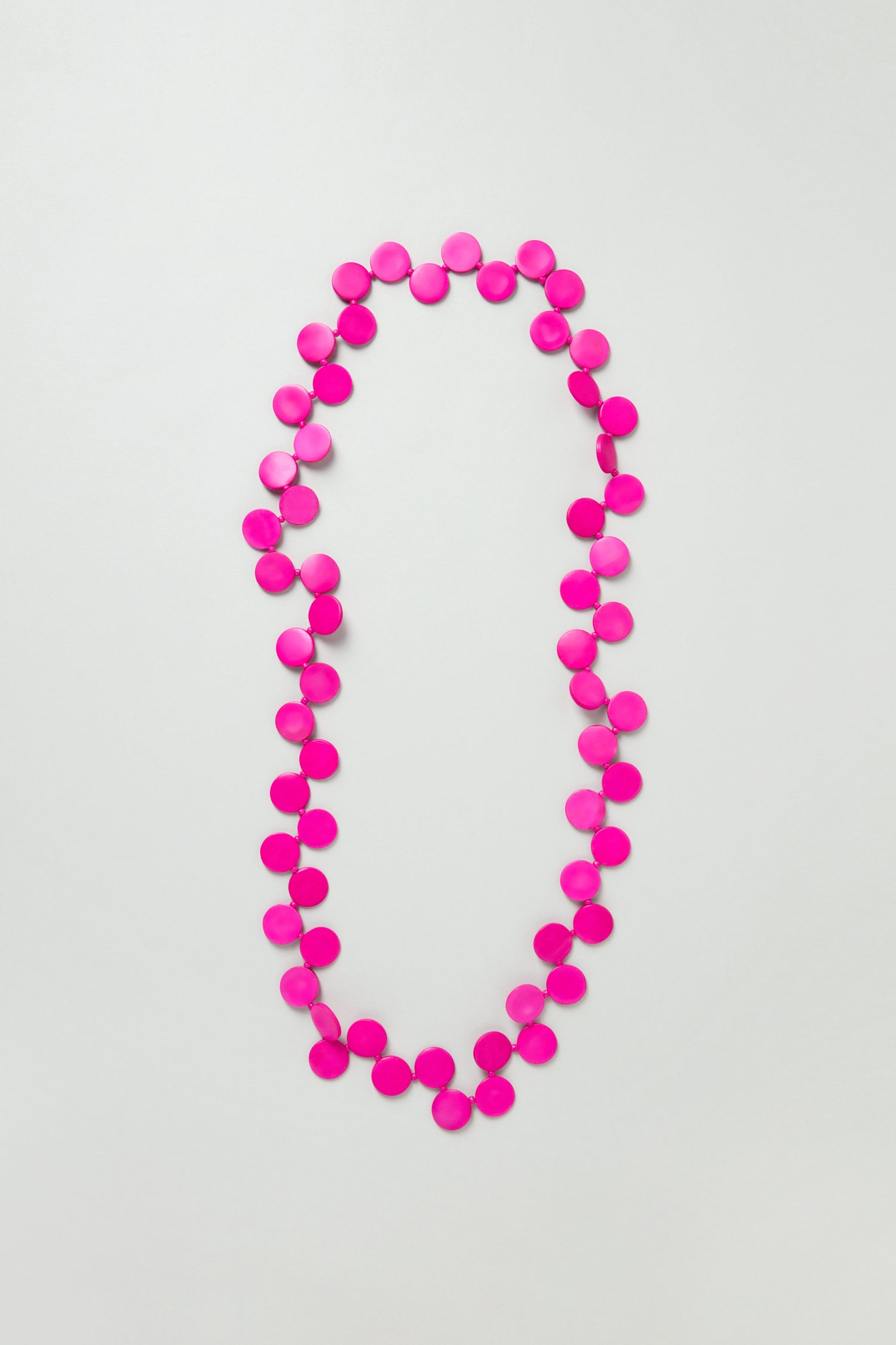 The Long Strand Coco Disc Necklace features handcrafted bright pink circular beads, arranged in a loose oval shape on a plain white background.