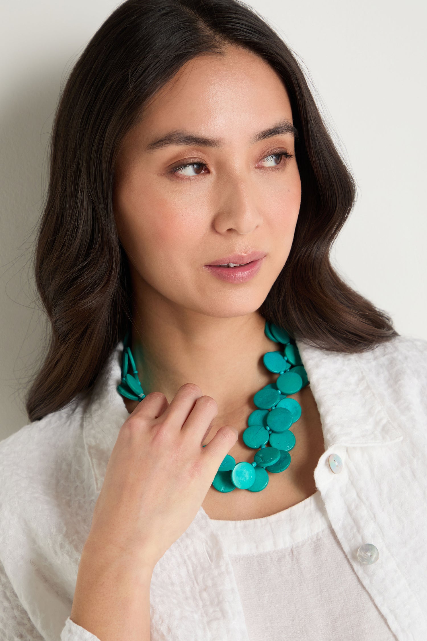 Woman with long dark hair wearing a white textured shirt and a Long Strand Coco Disc Necklace, looking slightly to the side.