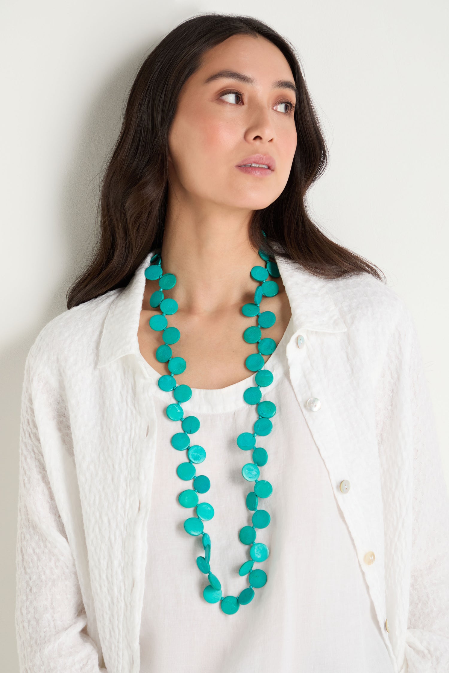 A woman stands against a light-colored wall wearing a white textured shirt and the Long Strand Coco Disc Necklace in turquoise.