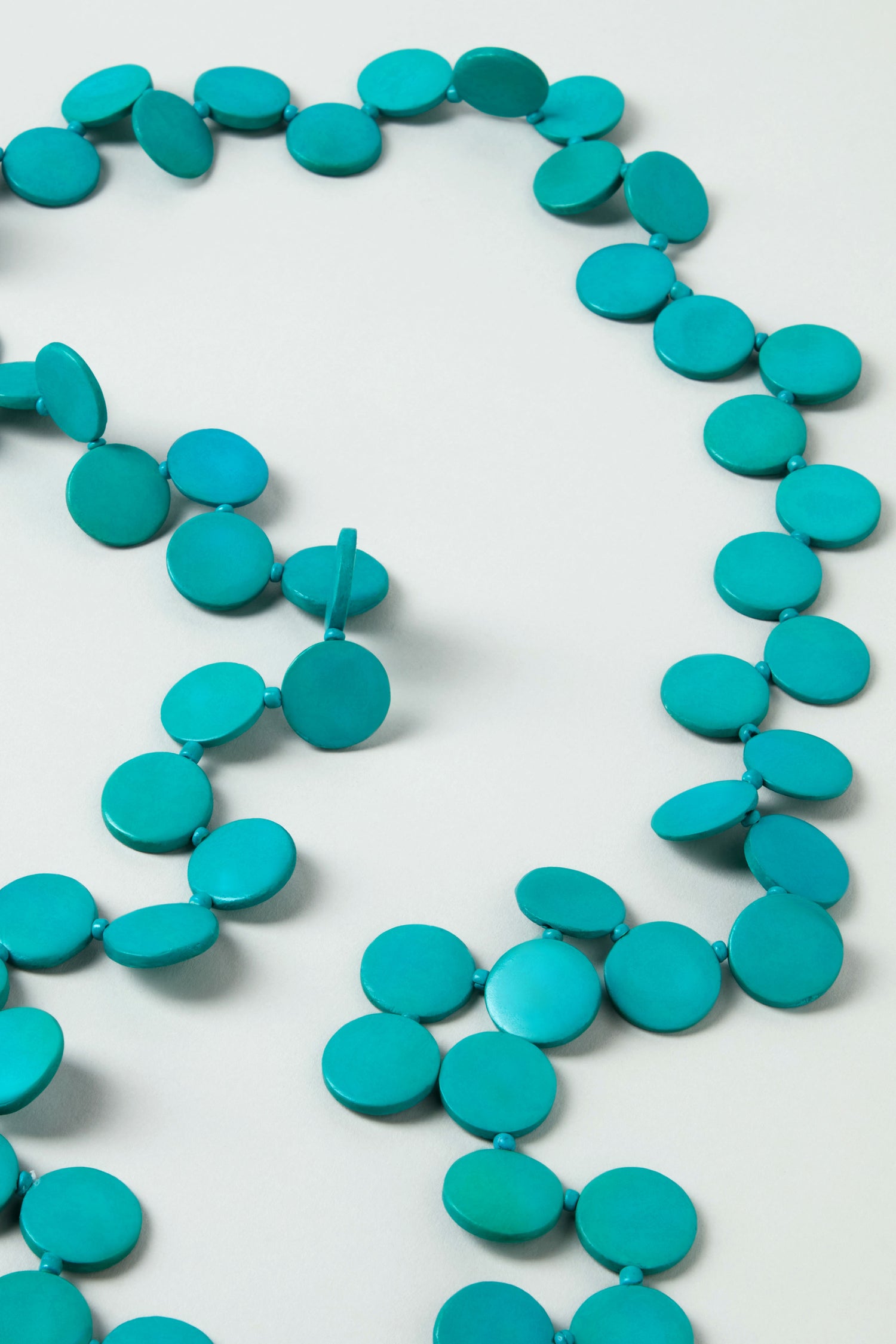 The Long Strand Coco Disc Necklace is handcrafted with round, flat turquoise beads arranged in a single strand and displayed on a light surface.