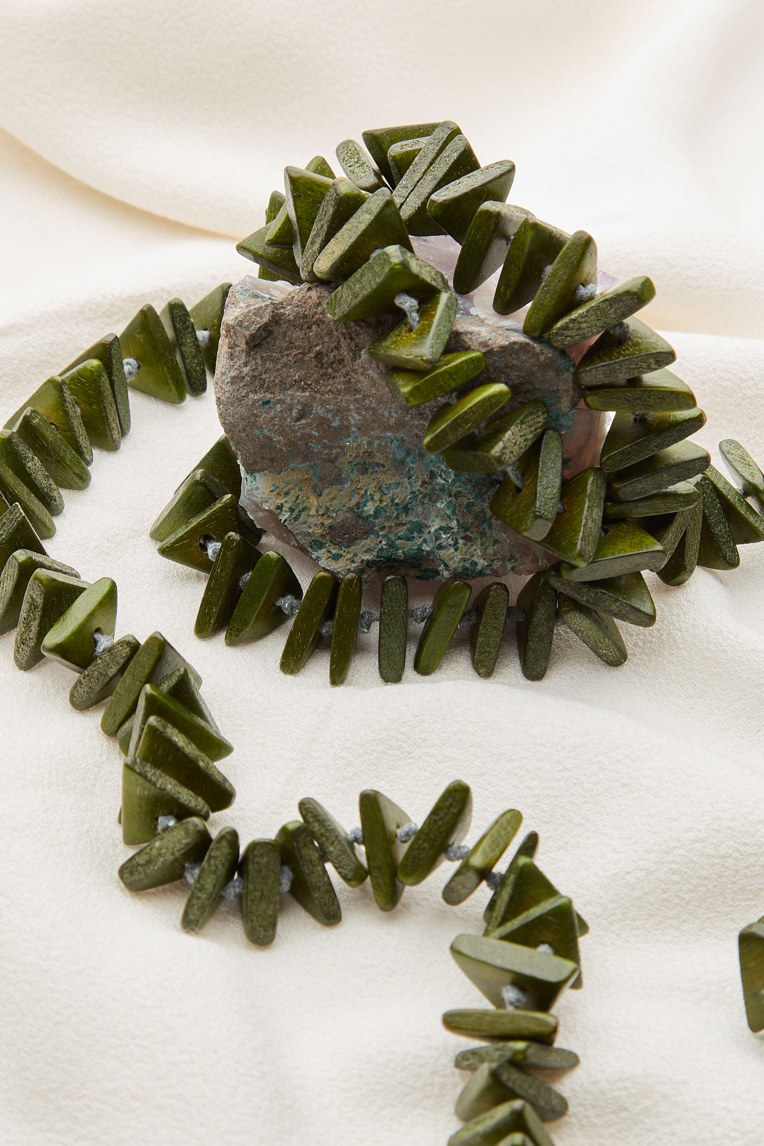 The Mini Triangles Wooden Necklace, adorned with green, triangular-shaped beads, is gracefully draped over a rock against a white fabric backdrop. This handmade wooden jewelry piece adds a touch of natural elegance to any setting.