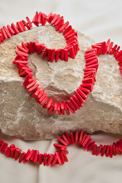 A Mini Triangles Wooden Necklace, featuring red, irregularly-shaped beads that mimic coral, is displayed over a textured rock on a light-colored surface, evoking the charm of handmade wooden jewelry.