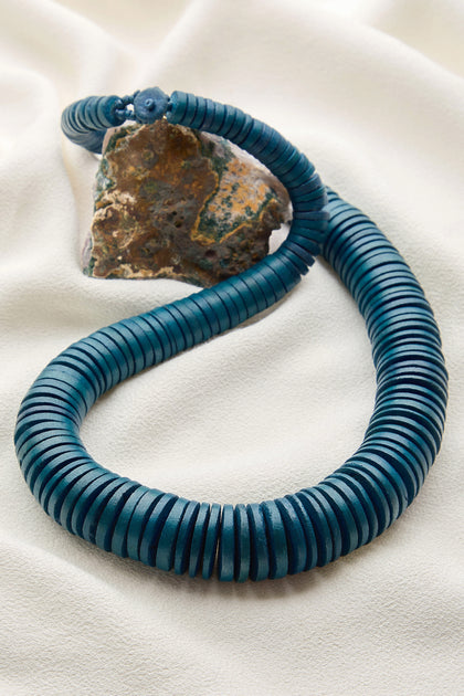 A Graduated Coco Disc Necklace, handmade in the Philippines with a coiled string of dark teal beads, rests on a light, textured fabric and features a rough, earthy stone at its end.