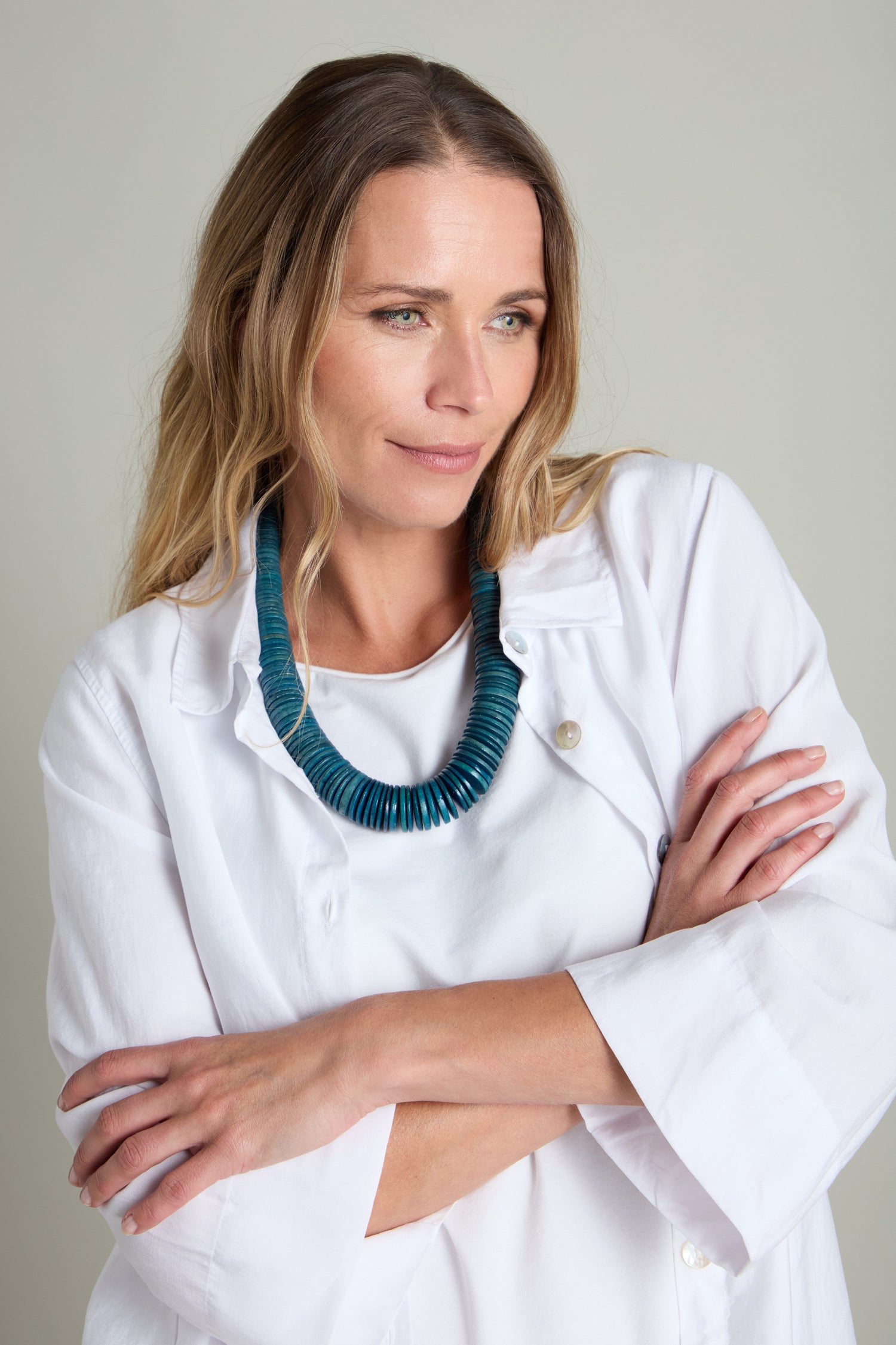 A person with long hair stands with arms crossed, wearing a white shirt and a blue Graduated Coco Disc Necklace, known for its unique shapes, as they look to the side.