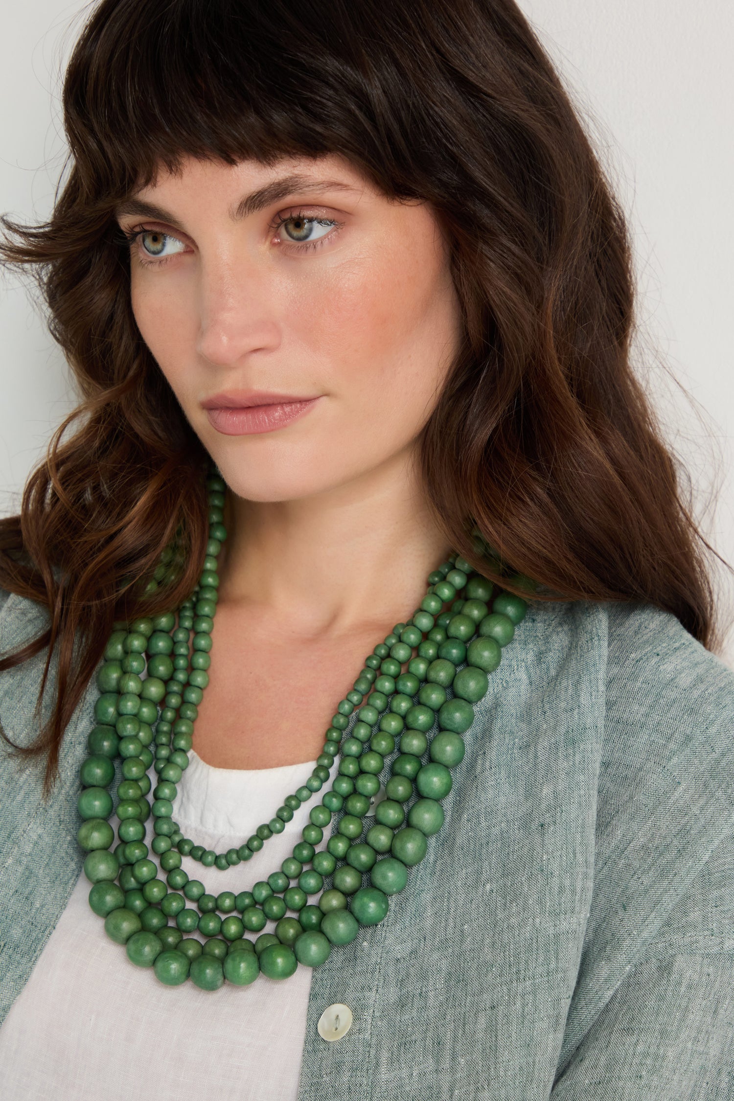 A woman with wavy brown hair wears a light green cardigan, white top, and the Statement Bead Necklace in green hues, looking slightly to the side.