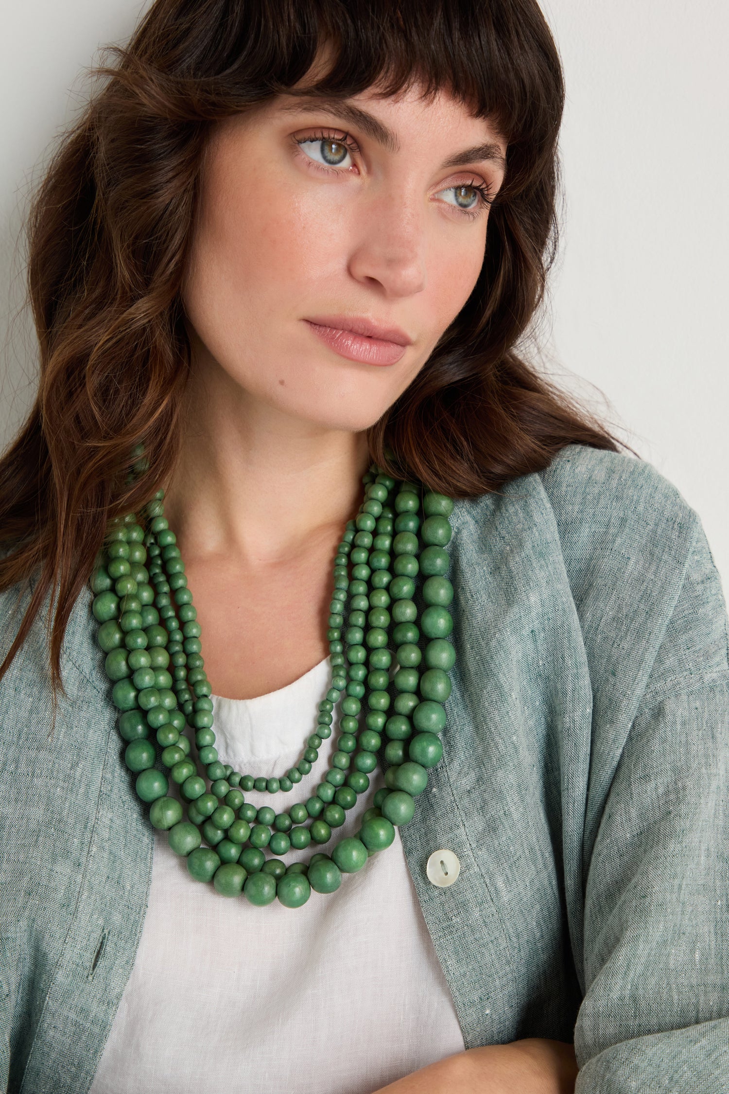 A woman with wavy brown hair wears a light green shirt, white top, and the Statement Bead Necklace—layered green sustainable wood beads—while gazing slightly to her right.