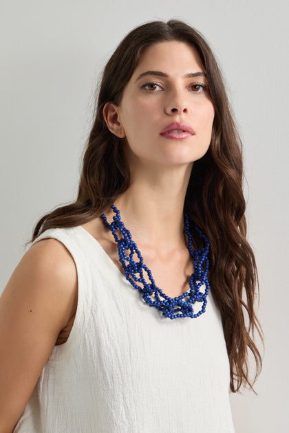 A long-haired woman in a cream sleeveless top showcases the artisan-crafted Woven Coco Sphere Necklace, featuring layered blue coconut beads, against a plain background.