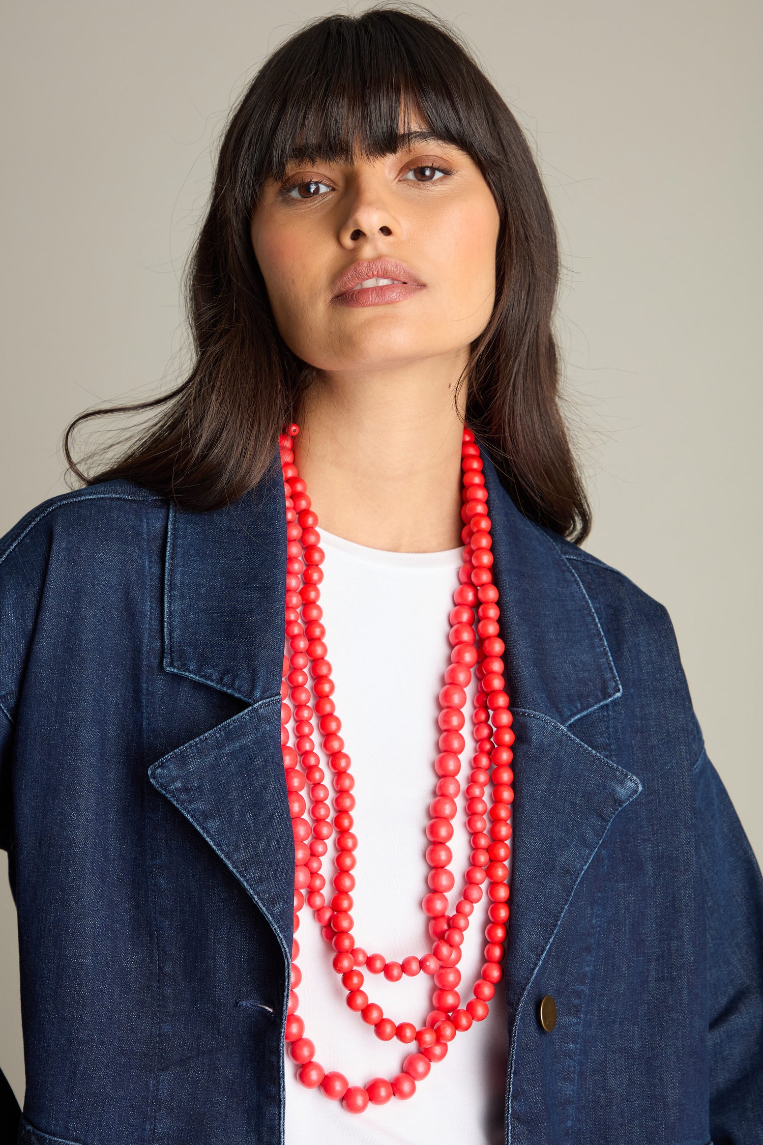 A woman with long dark hair wears a navy blue jacket, a white top, and a carefully handmade Wooden Cascade Spheres Necklace in vibrant orange.