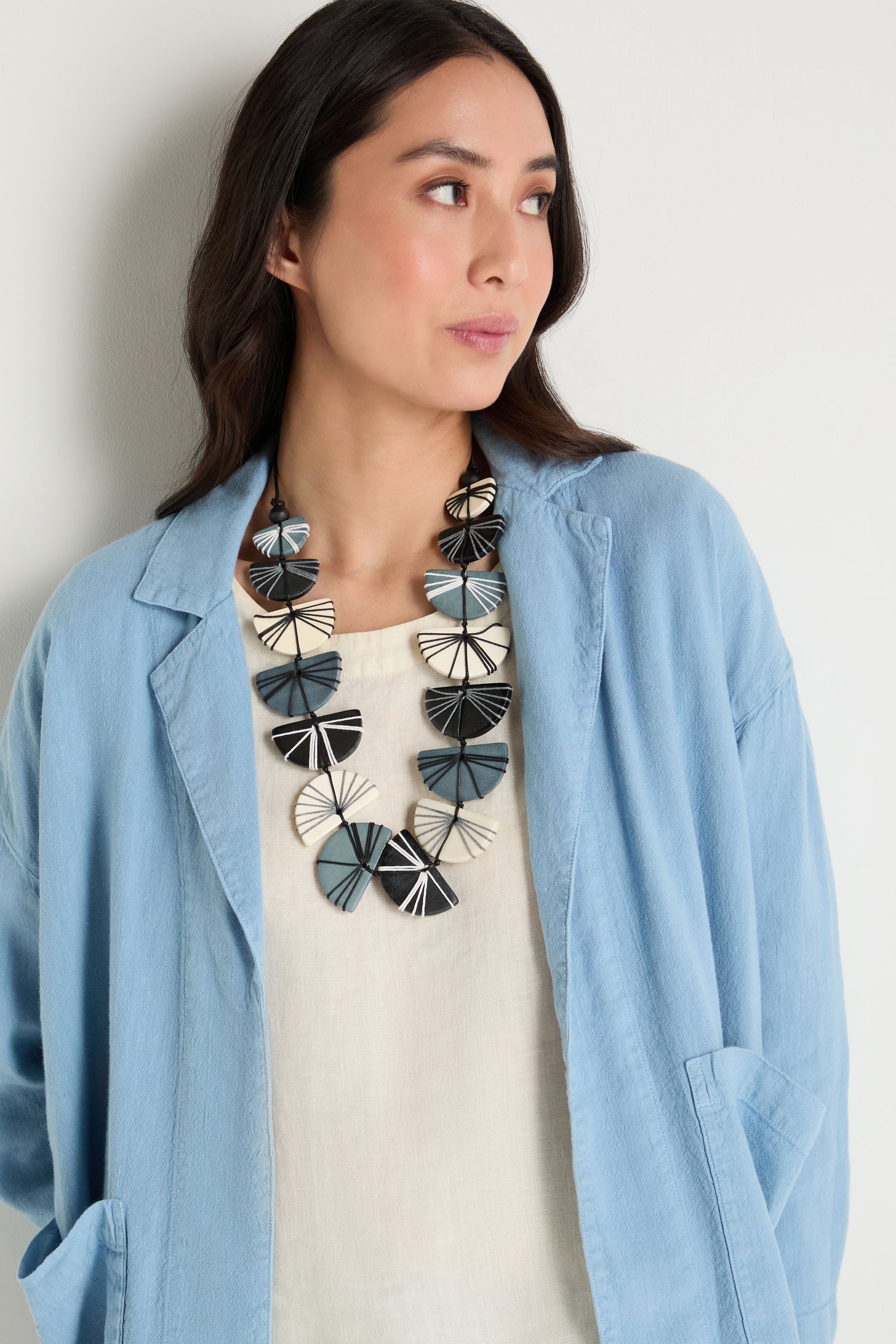 A woman in a light blue jacket and cream top wears the Fan Necklace, crafted from sustainable Pangantuan wood by Lotusfeet, while standing against a plain light-colored wall.
