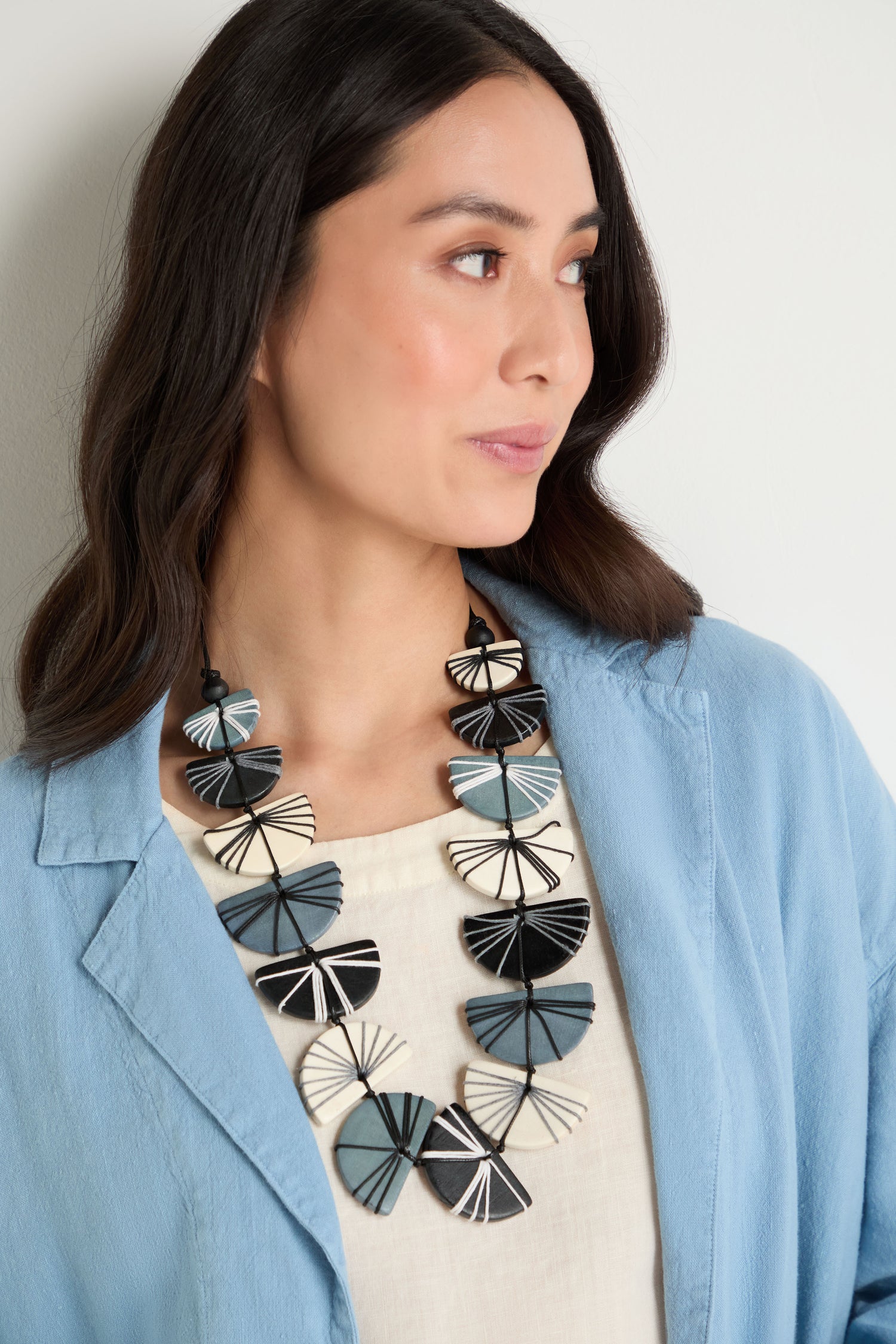 A woman in a light blue blazer and cream top wears the Fan Necklace, featuring bold black, white, and blue geometric shapes against a light background.