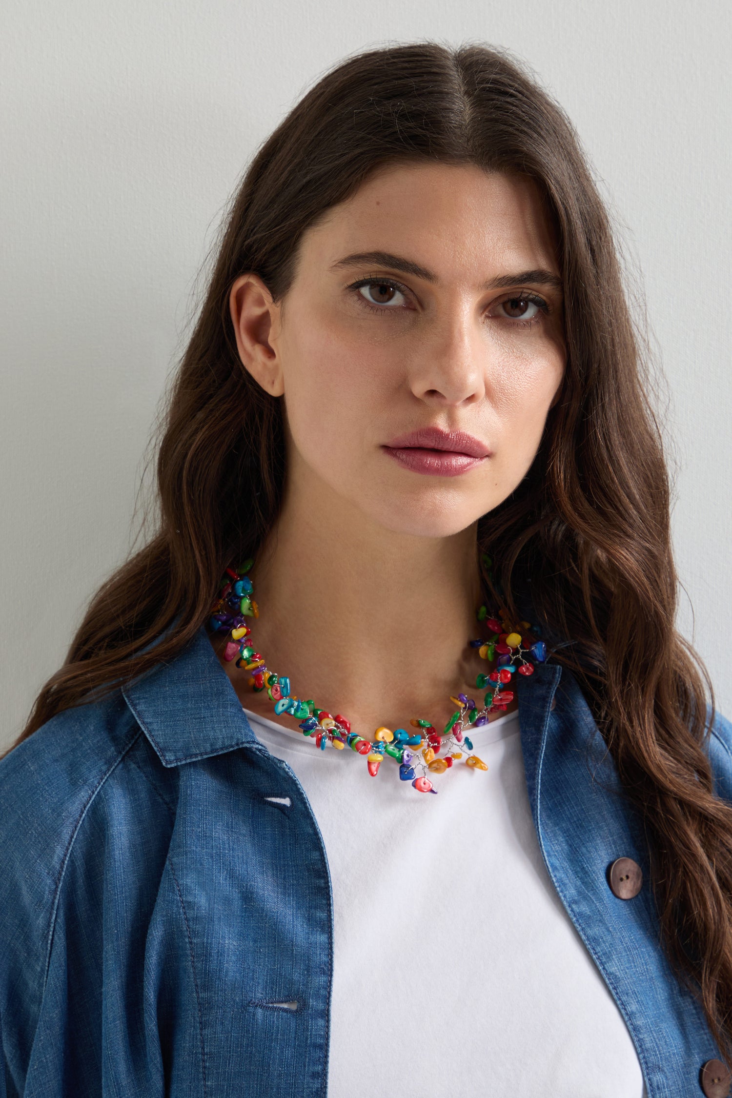 A woman with long dark hair in a blue shirt models the Rainbow Rivershell Necklace, its handcrafted river shell beads standing out against the plain white background, creating a true statement accessory.