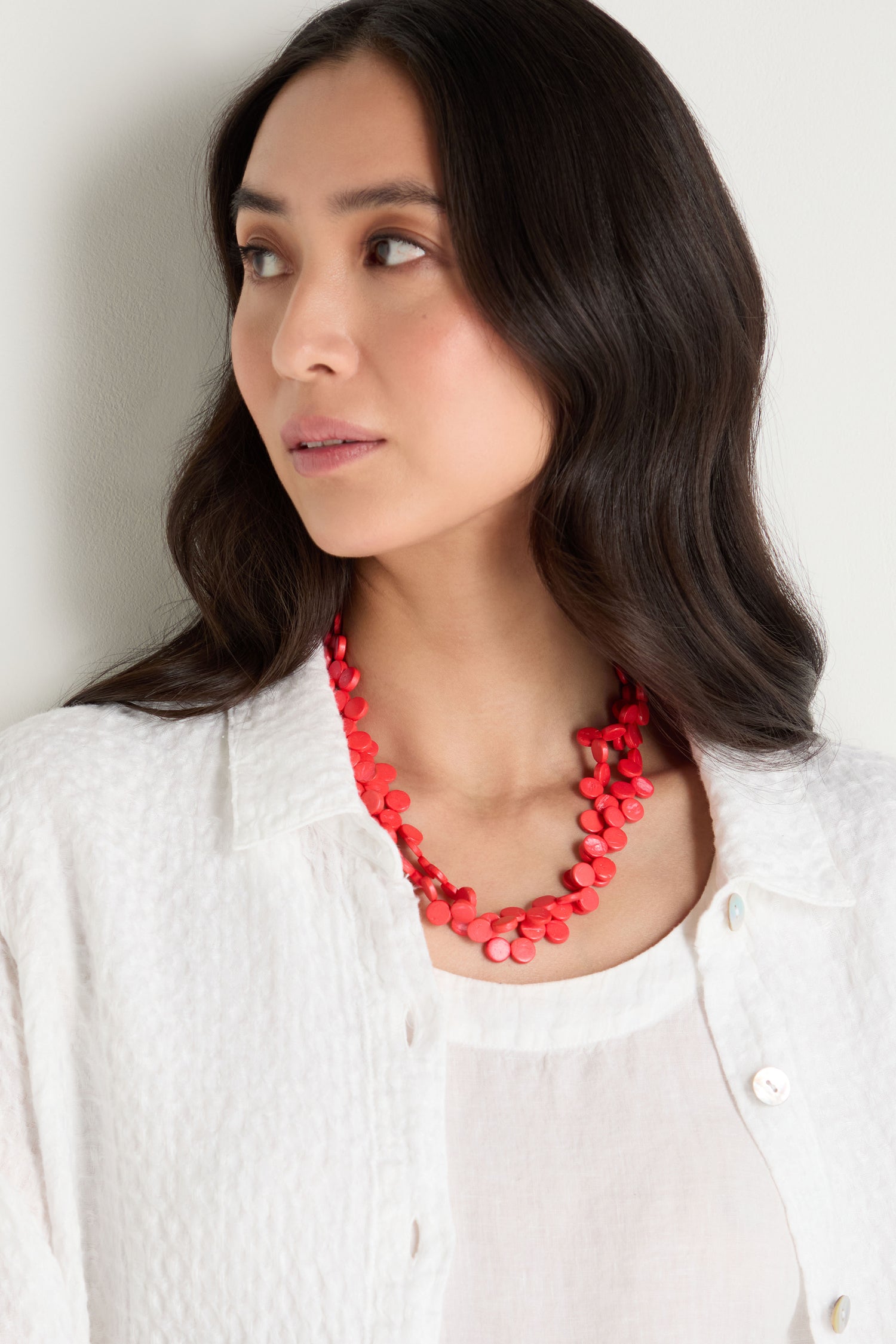 Woman with long dark hair wears a white textured top and the Single Strand Coco Necklace—a handcrafted red beaded accessory—while looking to the side against a light background.