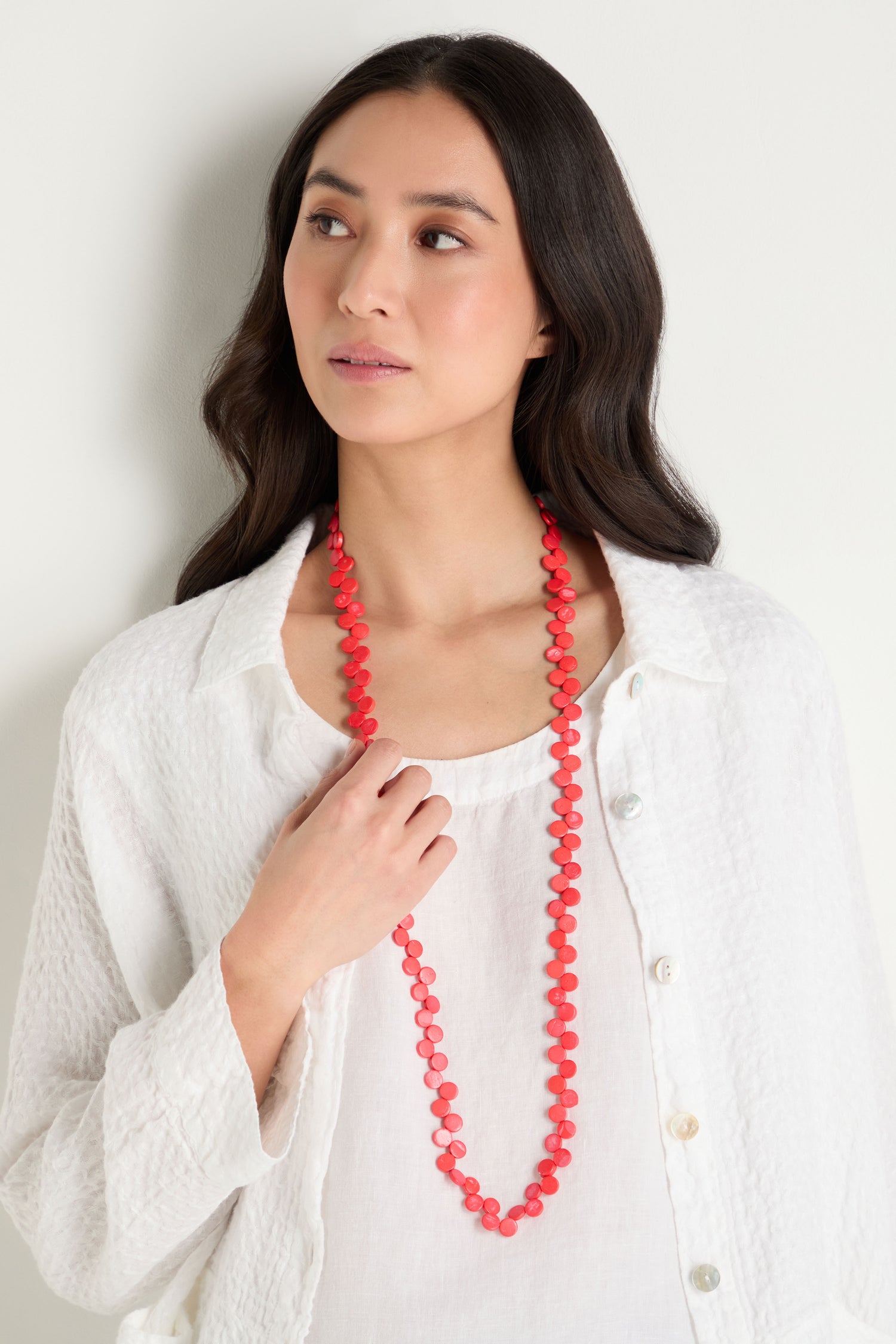 A woman with long dark hair wears a white textured shirt and the Single Strand Coco Necklace, looking slightly to the side against a plain background.