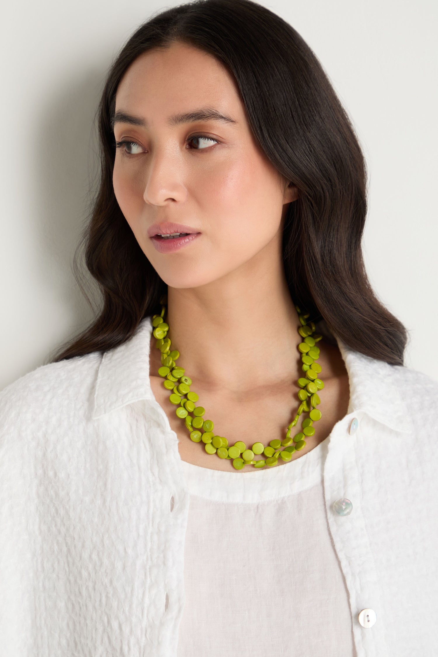 A woman with long dark hair, wearing a textured white shirt and the Single Strand Coco Necklace in vibrant lime green, looks slightly to the side against a light background.