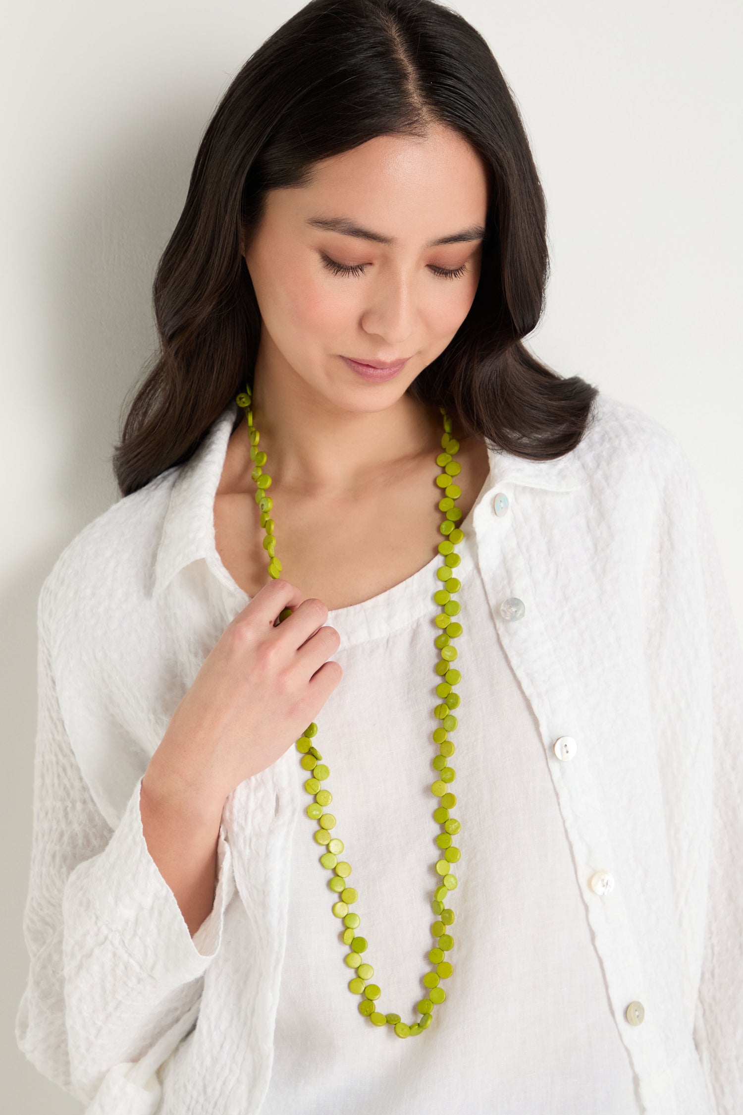 A woman in a textured white shirt stands against a white background, looking down while holding the Single Strand Coco Necklace—a long, bright green handcrafted accessory.