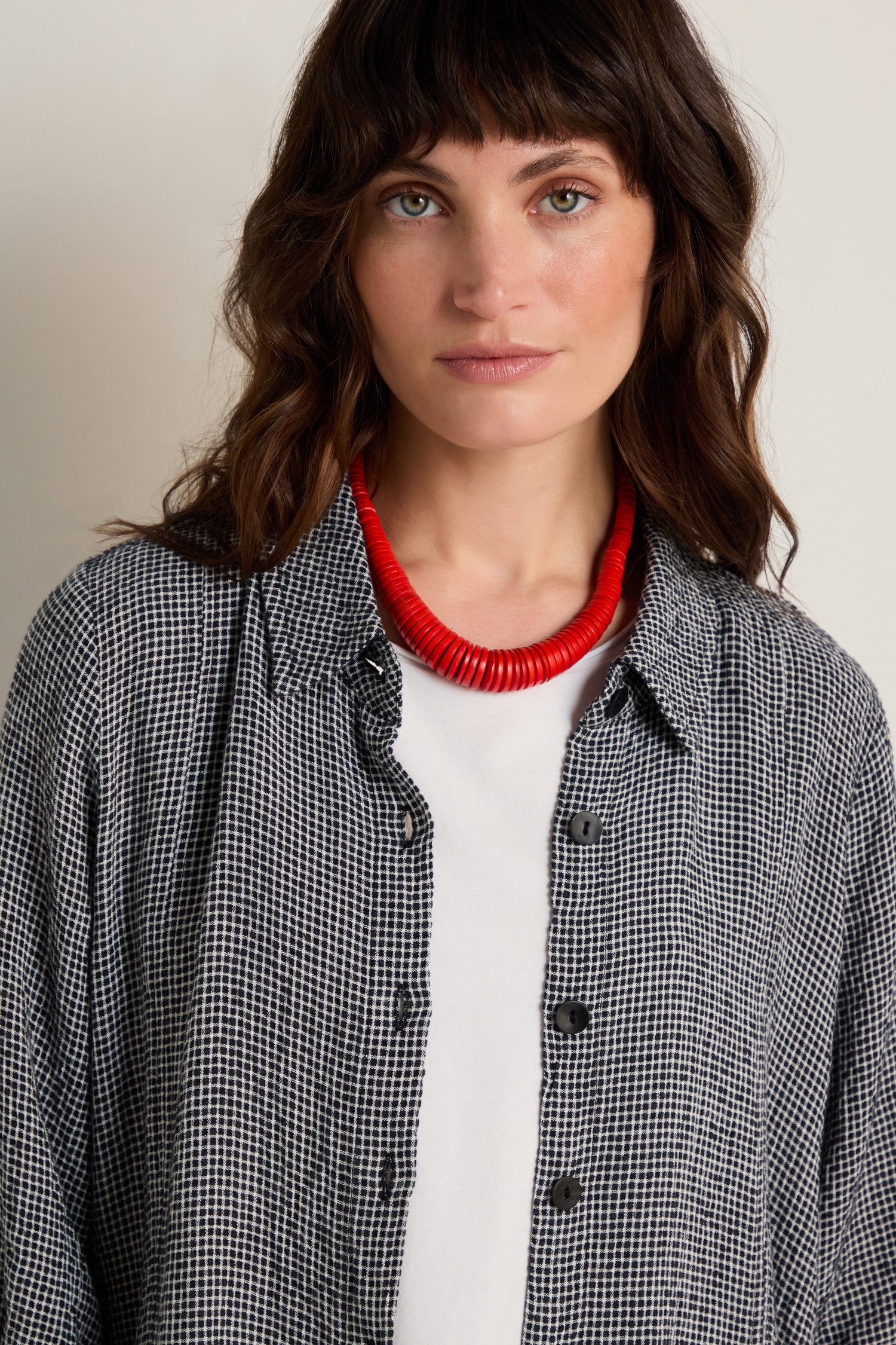 Woman with wavy brown hair wears a black and white checkered shirt, white top, and the Graduated Coco Disc Necklace—chunky red beads handcrafted in the Philippines—standing against a plain background.