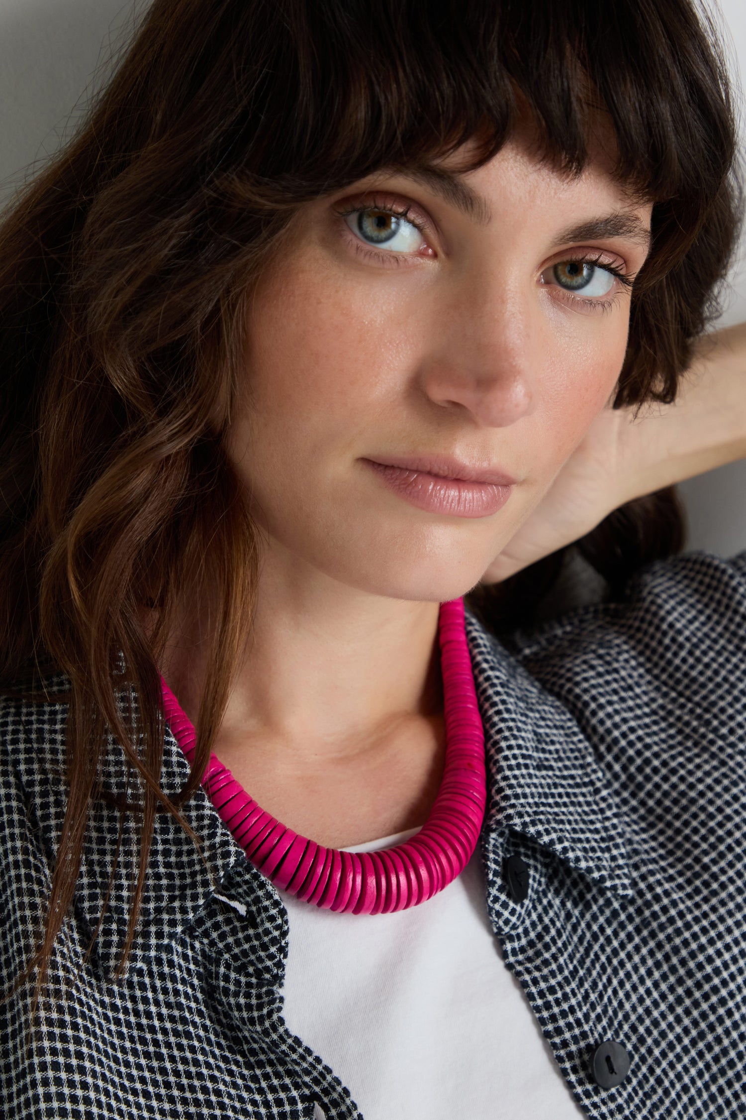 A woman with wavy brown hair wears a black-and-white checkered shirt, a white top, and a Graduated Coco Disc Necklace handcrafted and ethically sourced from the Philippines as she looks at the camera.