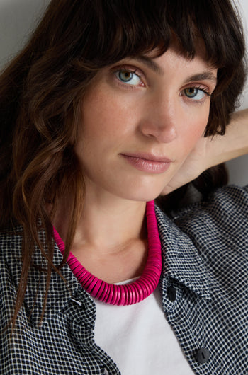 A woman with wavy brown hair wears a black-and-white checkered shirt, a white top, and a Graduated Coco Disc Necklace handcrafted and ethically sourced from the Philippines as she looks at the camera.
