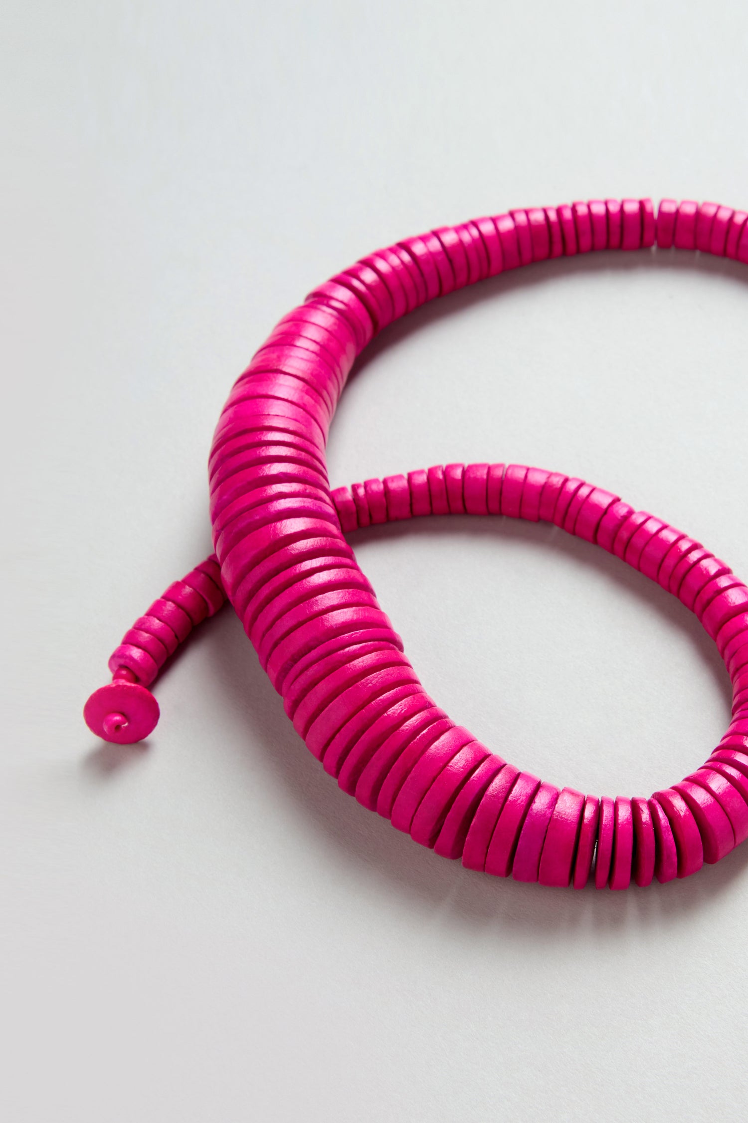 The Graduated Coco Disc Necklace features a coiled strand of bright pink, ethically sourced disc-shaped beads, beautifully arranged on a light gray background.