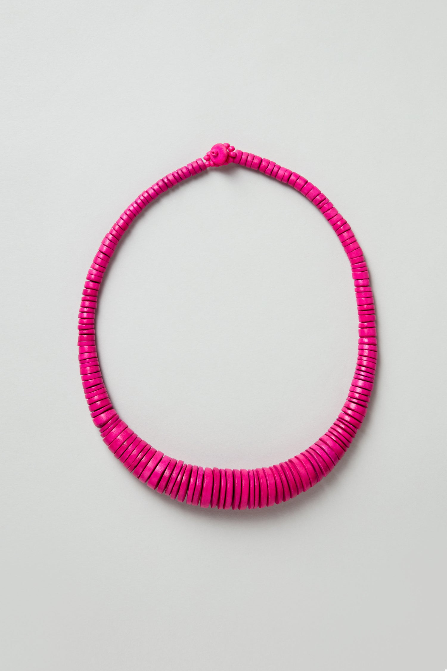 The Graduated Coco Disc Necklace is handcrafted in the Philippines, showcasing evenly spaced, round flat bright pink beads in a single strand, set against a light gray background.