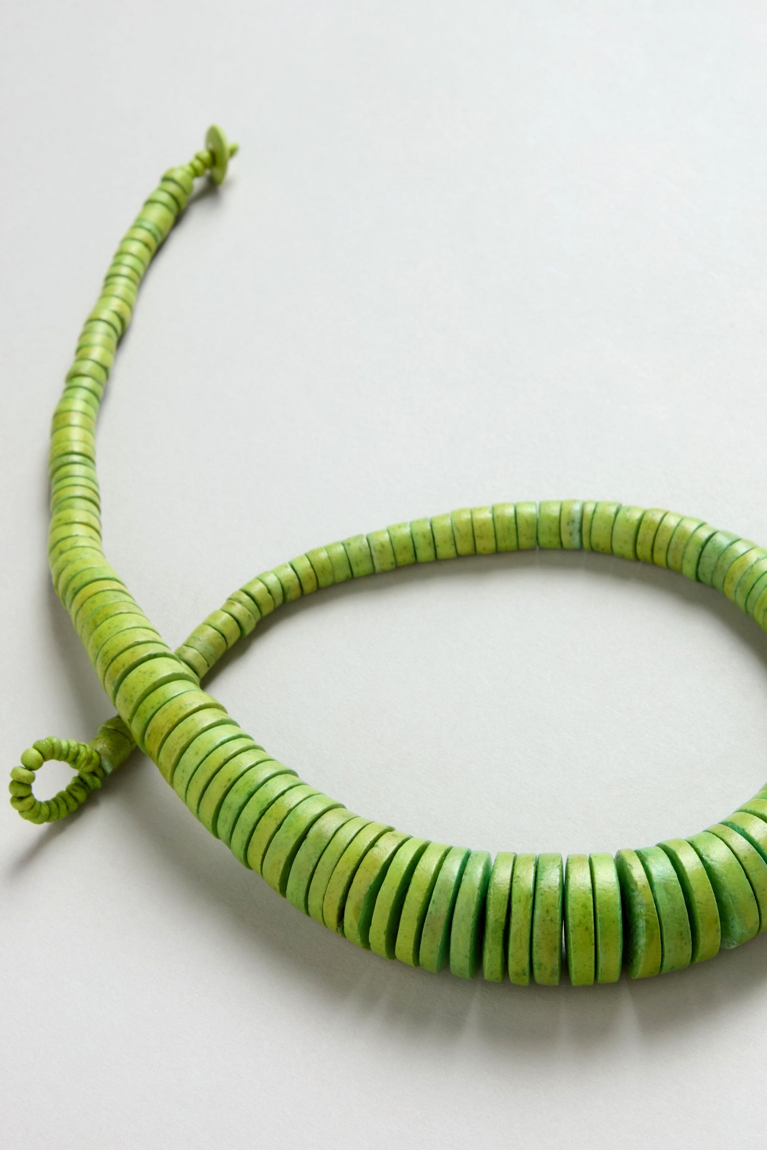 The Graduated Coco Disc Necklace, handcrafted in the Philippines from ethically sourced materials, features green graduated beads coiled on a light gray surface.