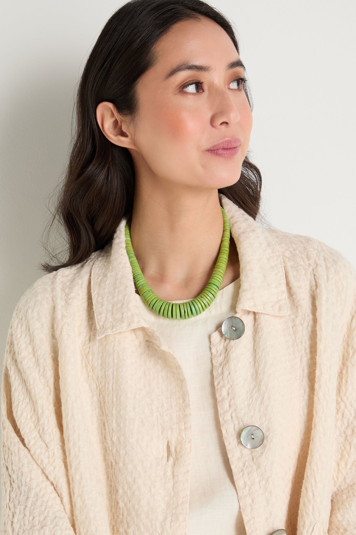 A woman in a textured cream jacket wears the Graduated Coco Disc Necklace, handcrafted in the Philippines, looking slightly to the side against a plain background.