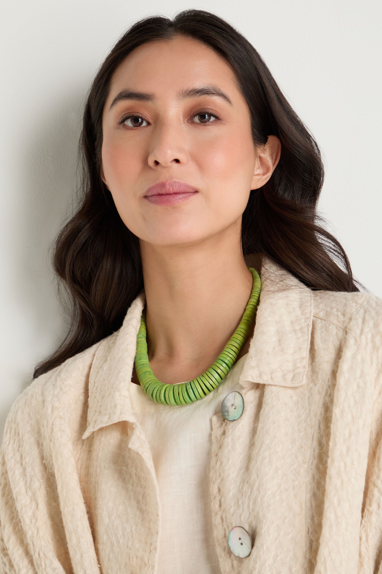 A woman with long dark hair wears the Graduated Coco Disc Necklace, handcrafted in the Philippines with ethically sourced materials, and a textured beige jacket, standing against a light background.