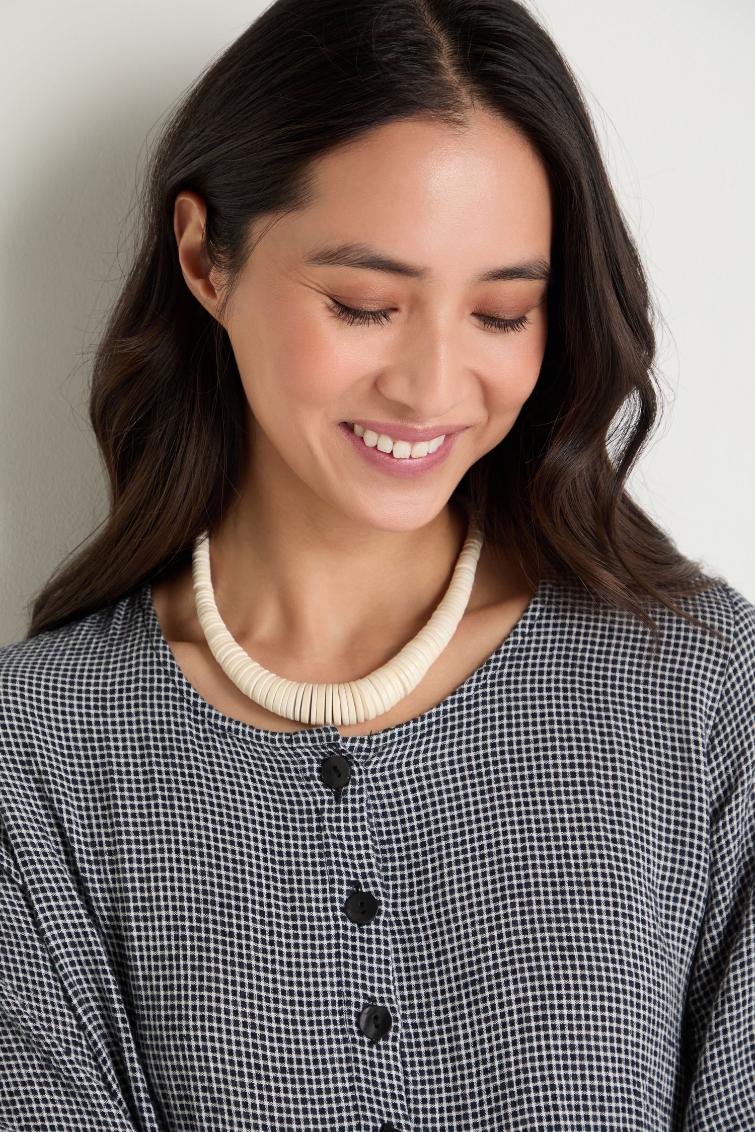 Woman with long dark hair, wearing a black and white checkered top and a Graduated Coco Disc Necklace, handcrafted in the Philippines, smiles while looking down.