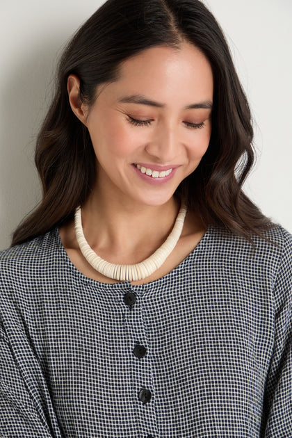 Woman with long dark hair, wearing a black and white checkered top and a Graduated Coco Disc Necklace, handcrafted in the Philippines, smiles while looking down.