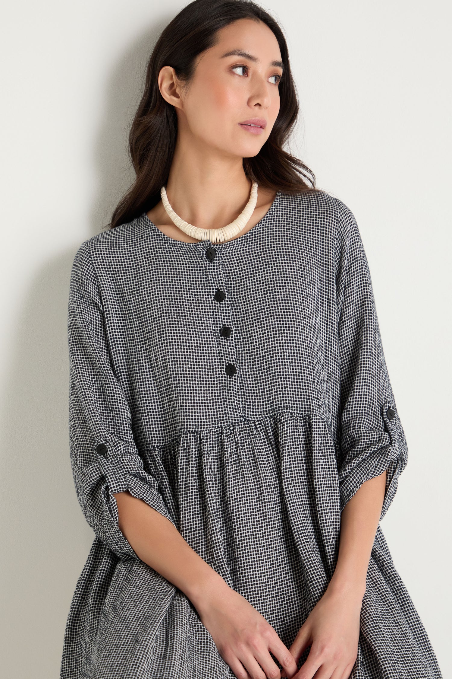 A woman in a black and white checkered dress with rolled-up sleeves wears the Graduated Coco Disc Necklace, handcrafted in the Philippines, as she stands against a plain light-colored wall.