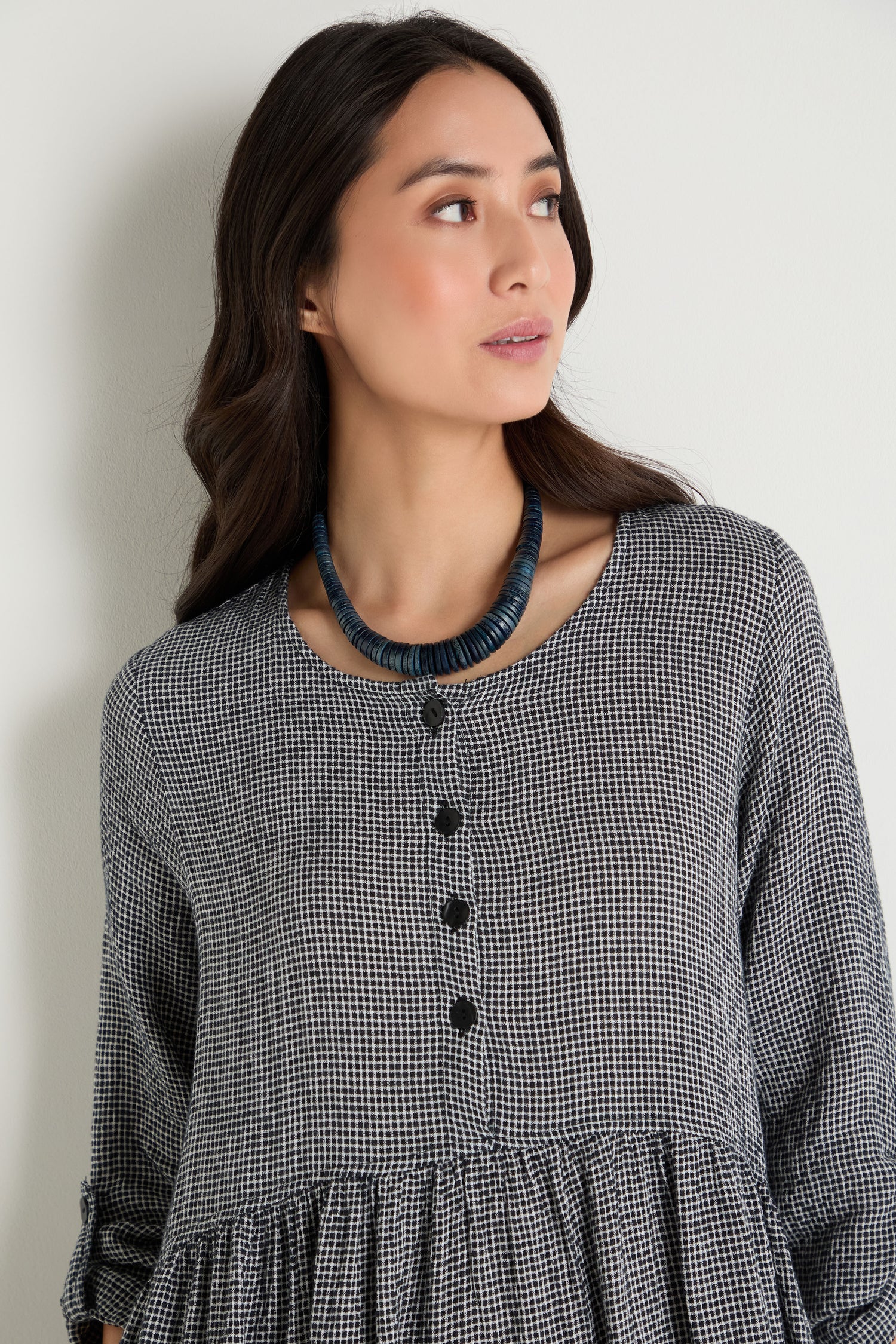 A woman with long dark hair wears a black and white checkered dress and the Graduated Coco Disc Necklace, ethically handcrafted in the Philippines, as she looks to the side against a plain light background.