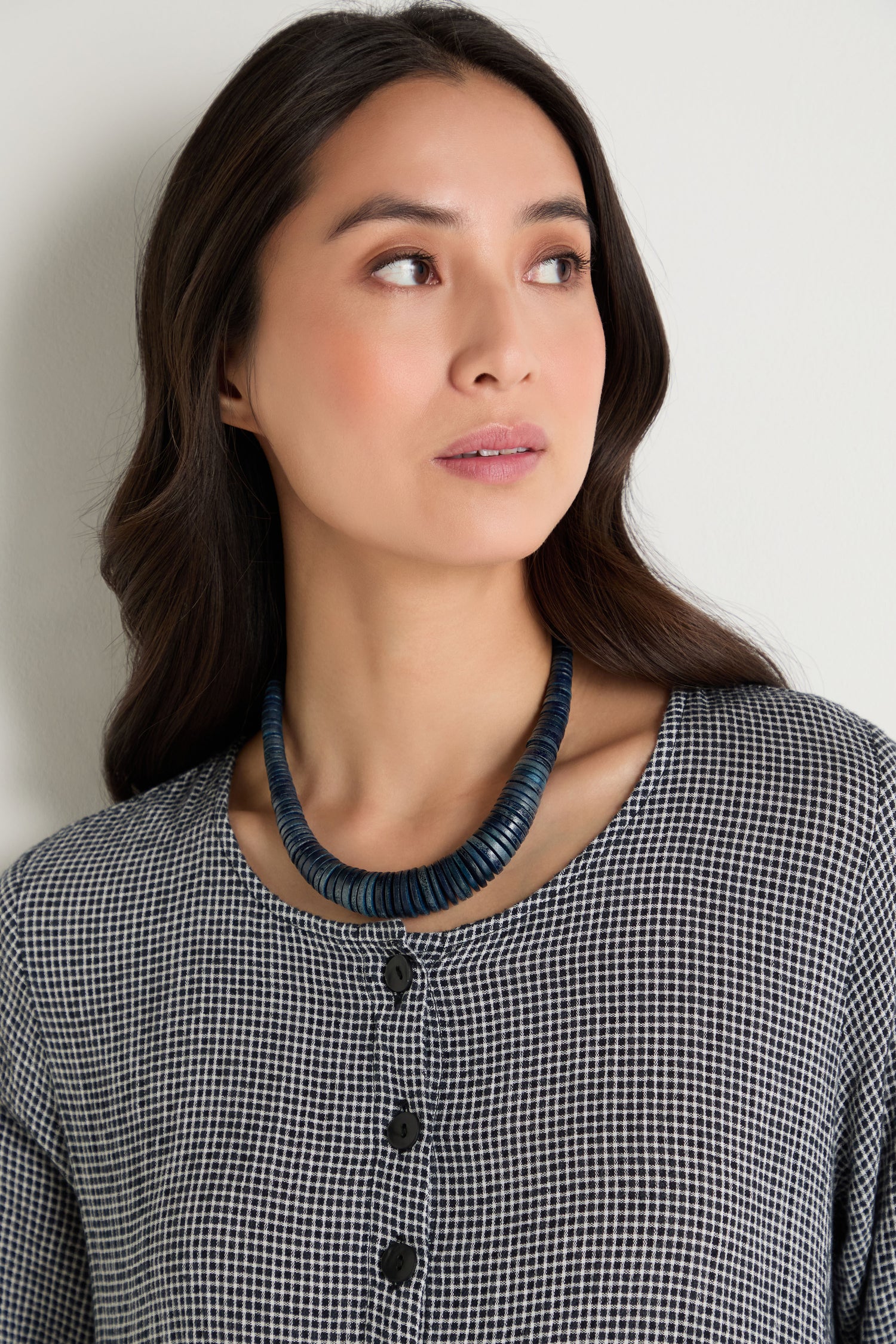 Woman with long dark hair wears a black and white checkered top and the Graduated Coco Disc Necklace, an ethically sourced chunky dark blue bead accessory handcrafted in the Philippines, as she looks to her left.