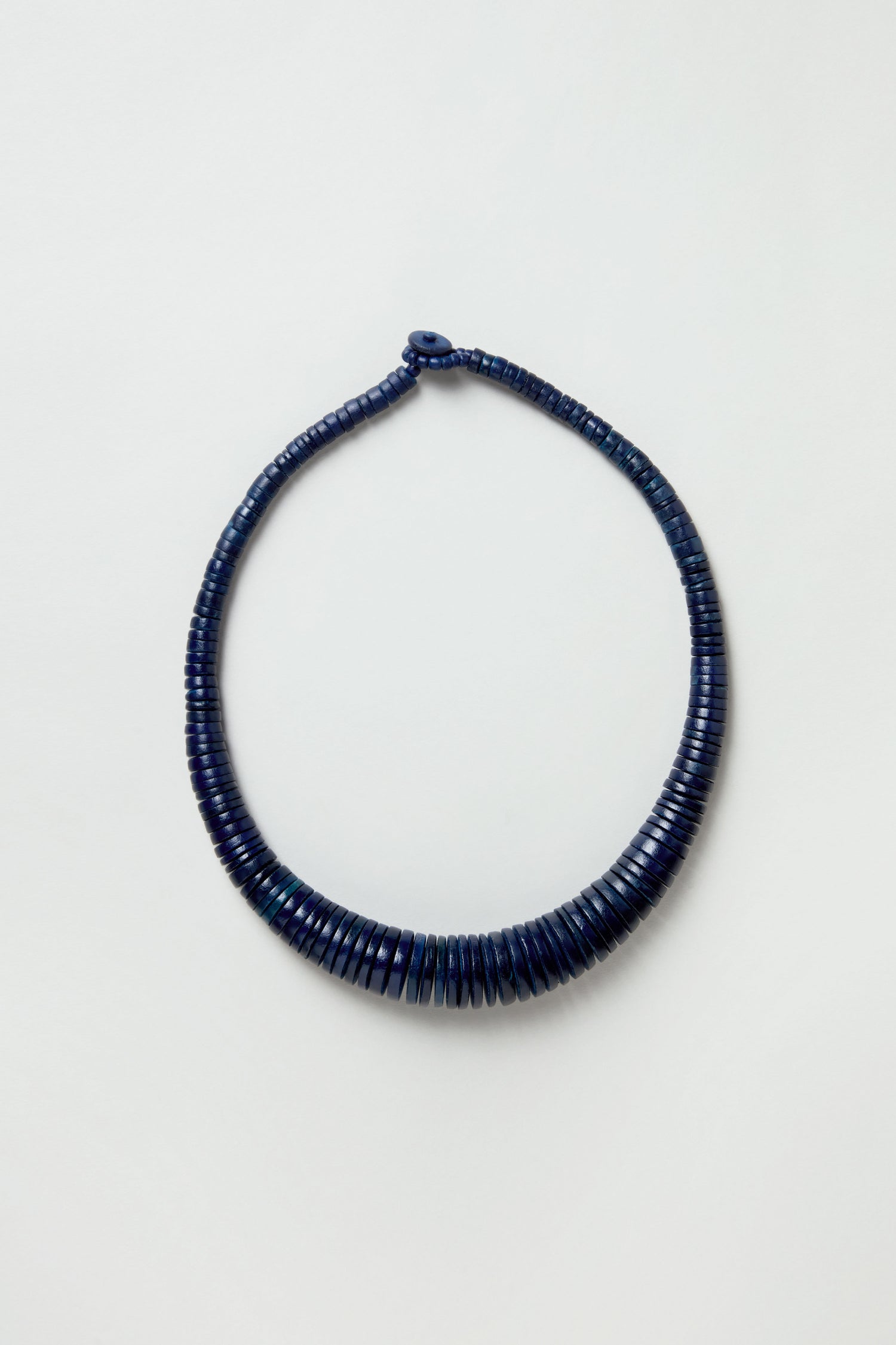 The Graduated Coco Disc Necklace is a closed-loop piece handcrafted in the Philippines, featuring uniformly stacked dark blue discs set against a light background.