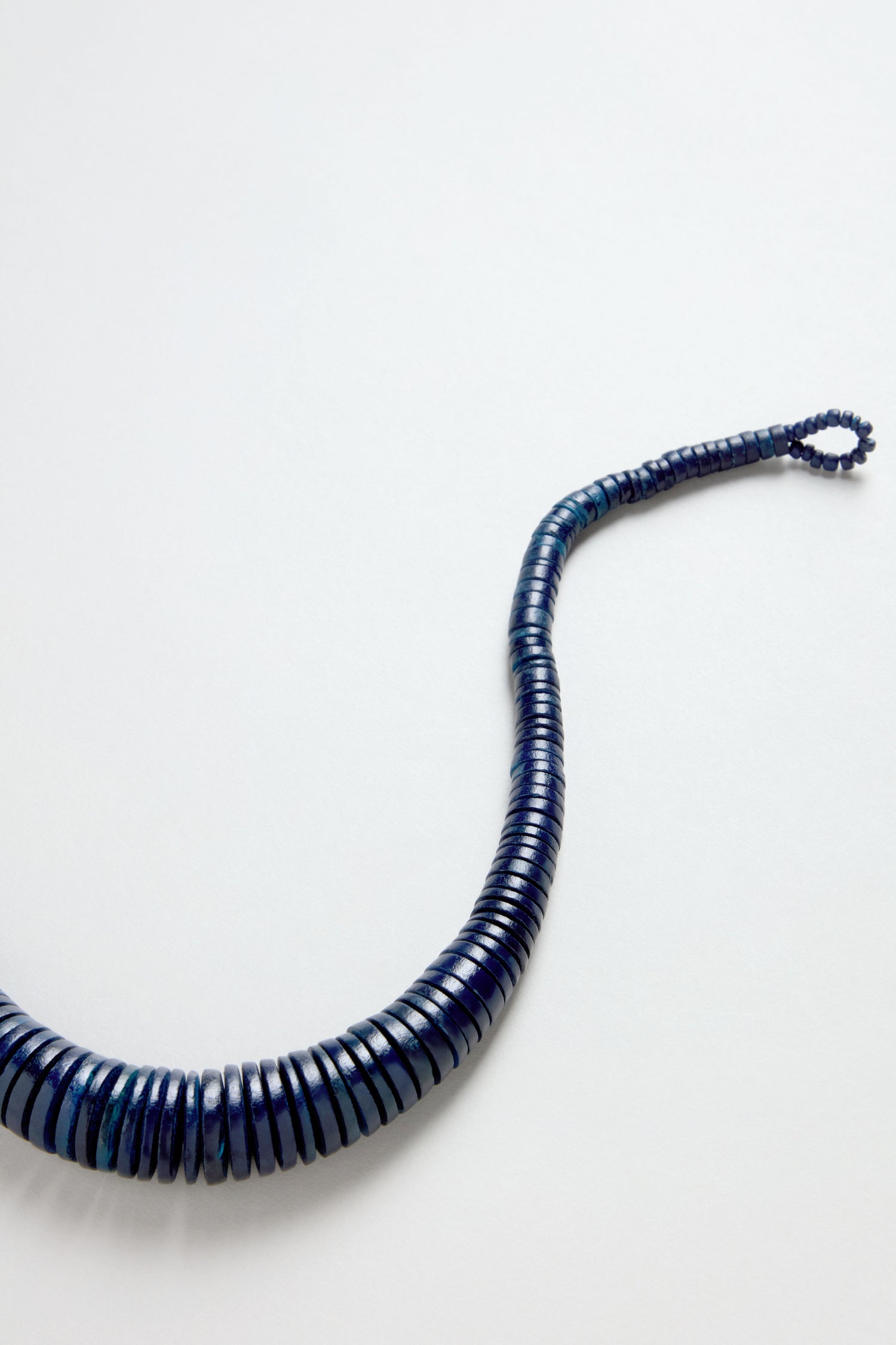 The Graduated Coco Disc Necklace, handcrafted in the Philippines, features coiled, segmented dark blue beads arranged in a graduated design and is displayed on a plain white surface, resembling a flexible beaded cord.