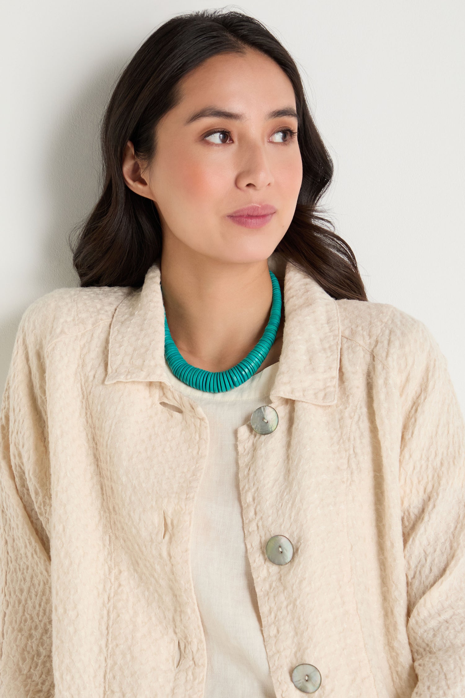 A woman with long dark hair, in a cream textured jacket and the Graduated Coco Disc Necklace—handcrafted in the Philippines from ethically sourced materials—stands against a light wall, gazing to the side.