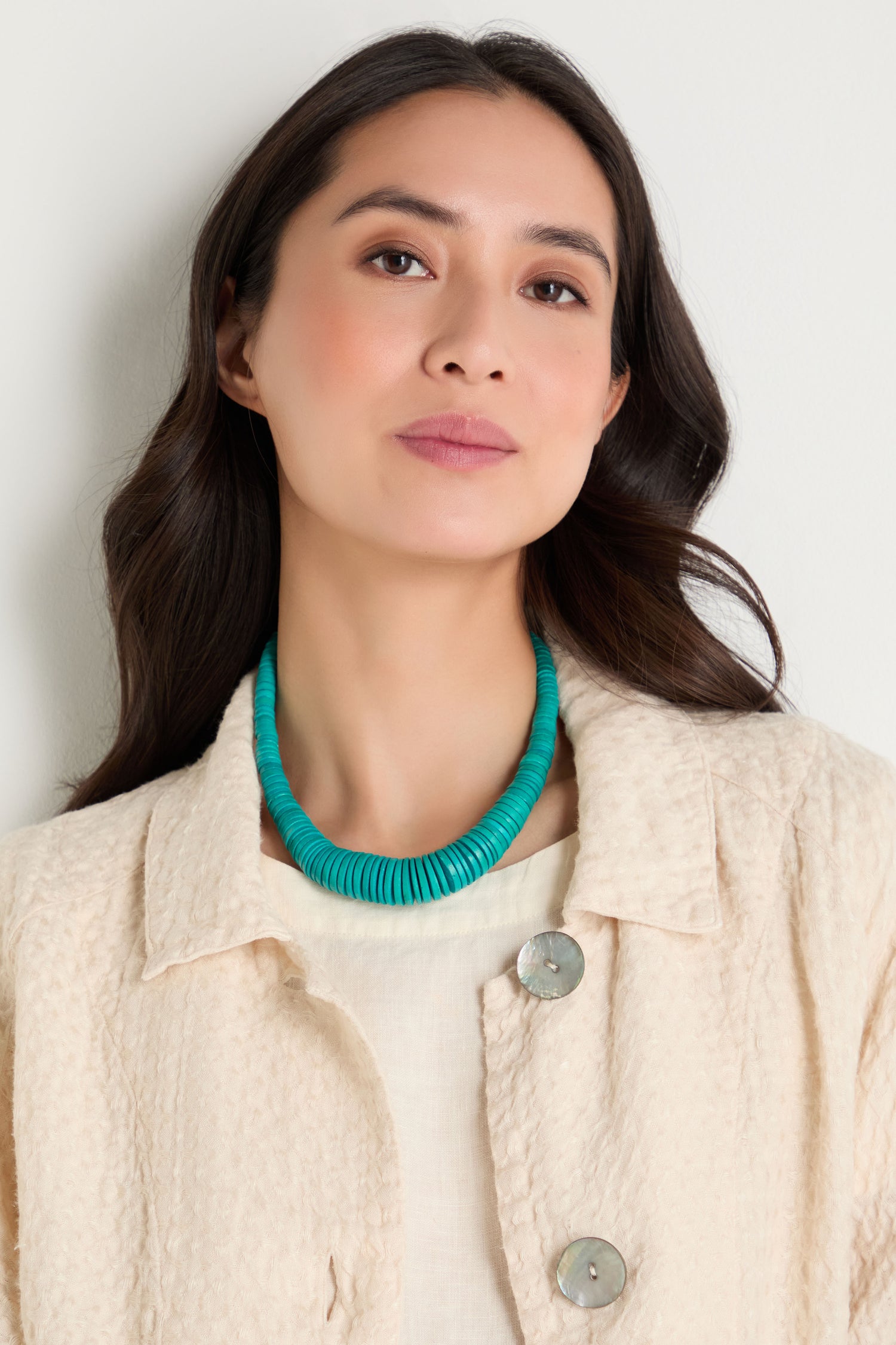 A woman with long dark hair wears a textured cream jacket and the Graduated Coco Disc Necklace, ethically handcrafted in the Philippines, standing against a plain, light-colored background.