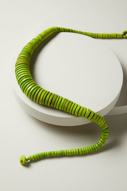 A vibrant green Graduated Coco Disc Necklace is displayed on a round white surface, showcasing its circular, disc-like beads and extending into a slightly curved shape. This ethically sourced piece boasts a bold hue that captures attention.