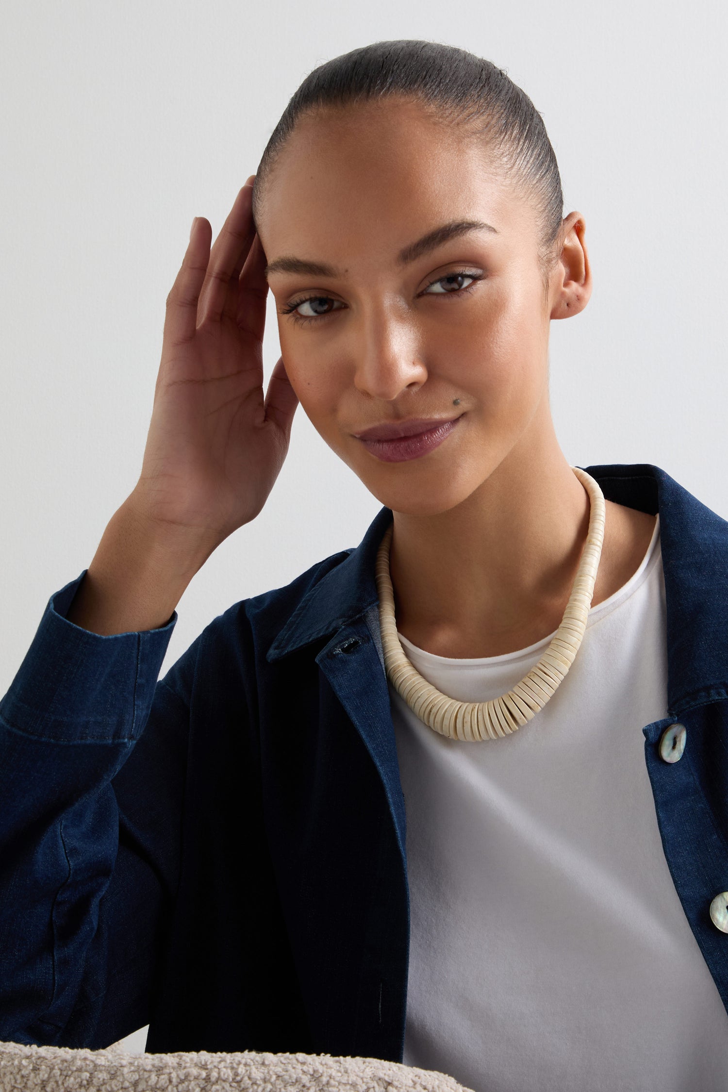 A person in a dark blue shirt wears the elegantly crafted Graduated Coco Disc Necklace in beige, seated with one hand on their head, smiling. The necklace showcases exquisite Philippine craftsmanship and adds an elegant touch to their cheerful presence.